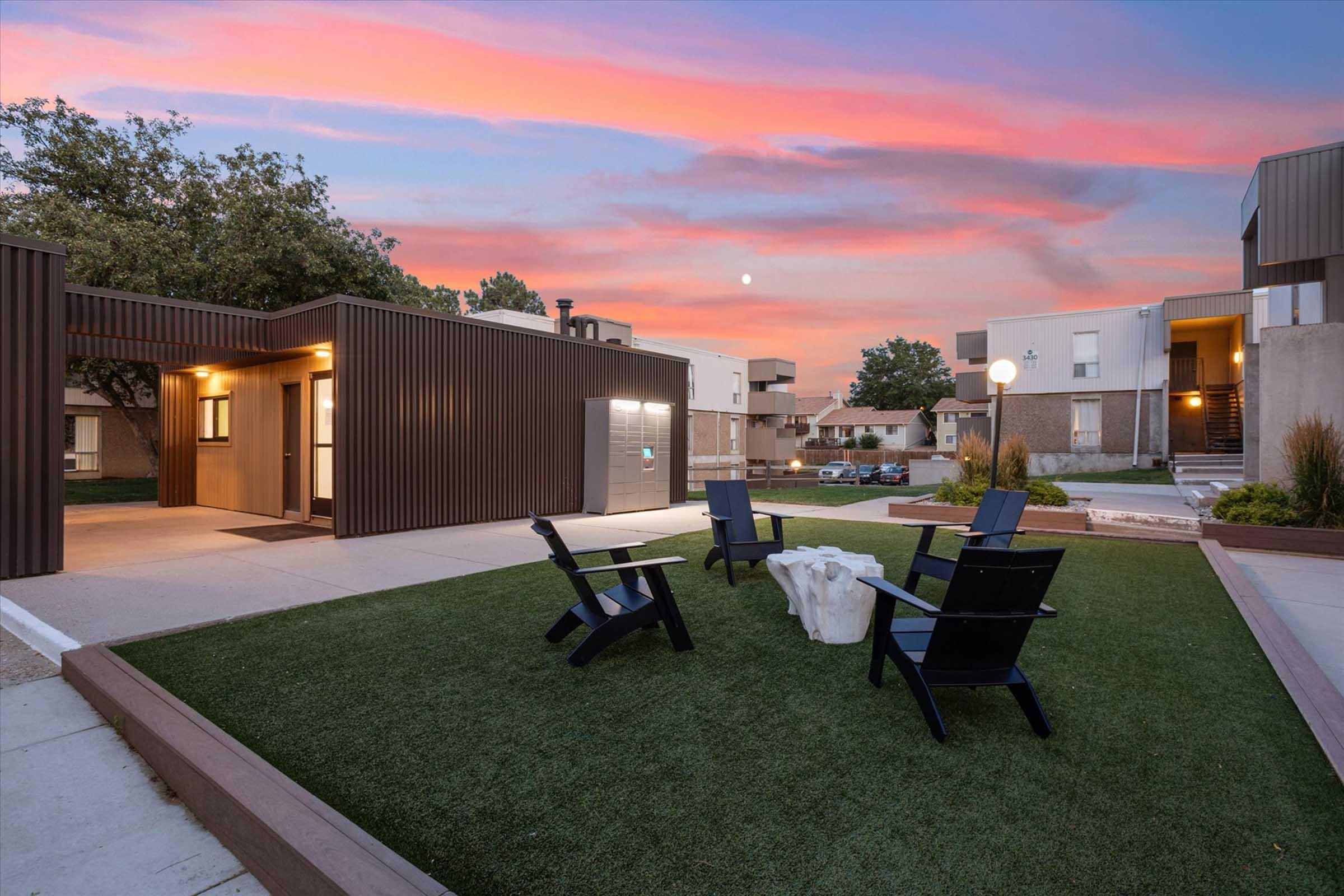 A modern outdoor area featuring a landscaped green lawn with several black Adirondack chairs arranged around a white stone fire pit. In the background, contemporary buildings are set against a colorful sunset sky with hints of pink and purple, and a full moon is visible.