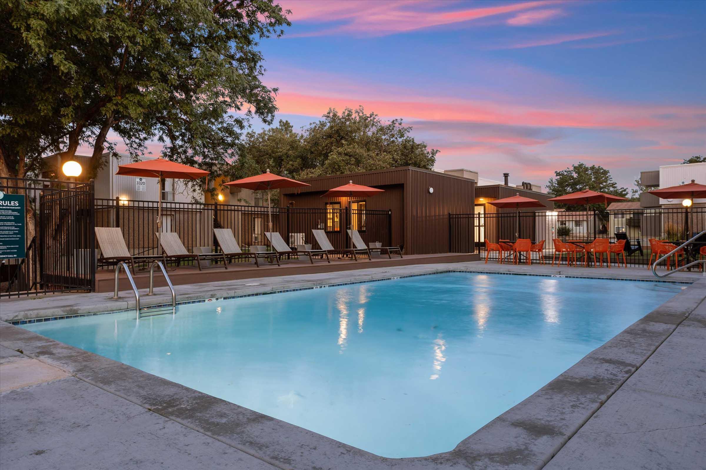 A serene swimming pool surrounded by lounge chairs and orange umbrellas, set against a backdrop of trees and a colorful sunset sky. The area is enclosed by a metal fence, providing a relaxed atmosphere for leisure and enjoyment.