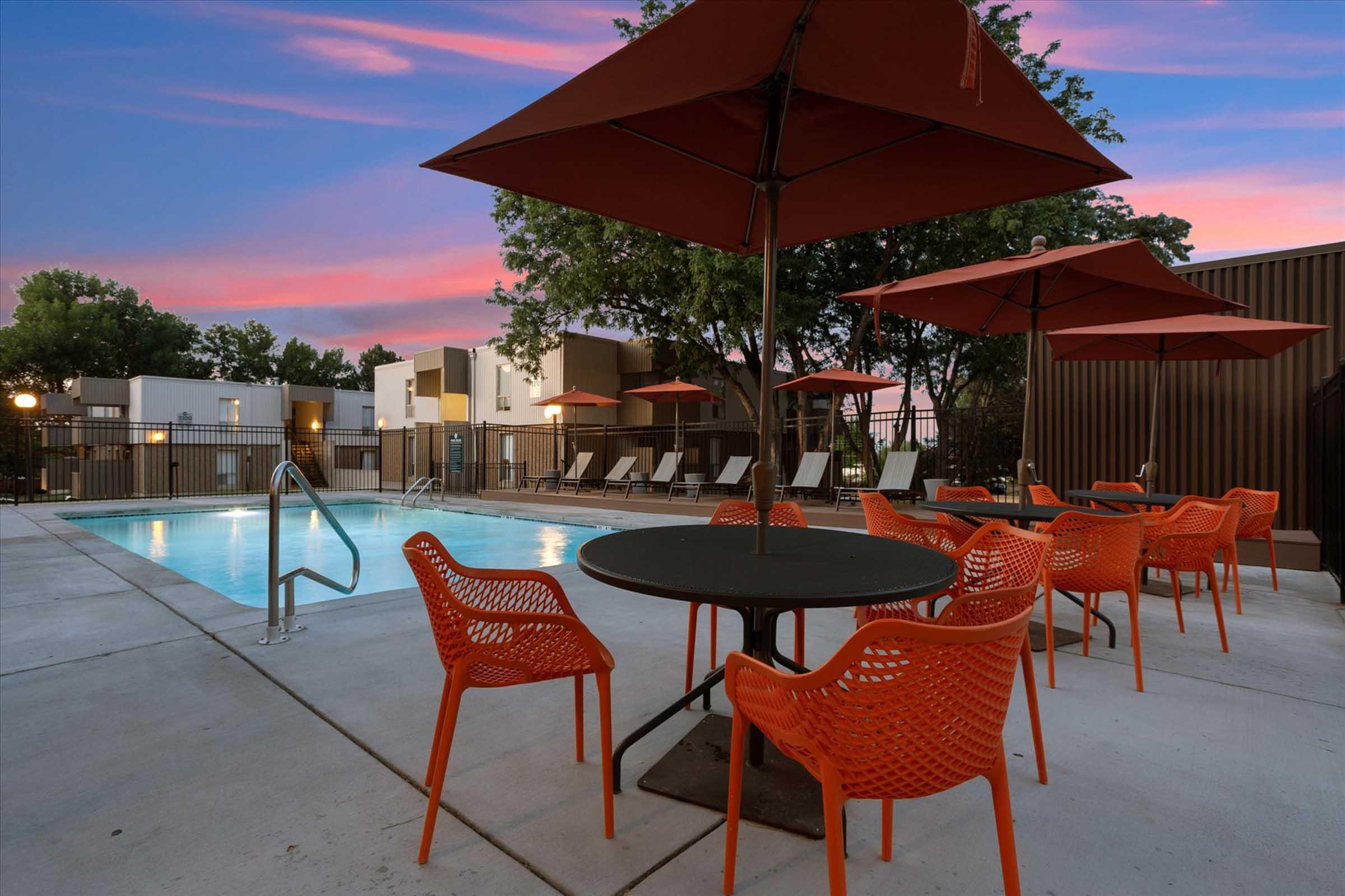 Outdoor lounge area featuring a swimming pool surrounded by orange patio furniture and umbrellas. The sky is painted in shades of pink and purple during sunset, with several lounge chairs adjacent to the pool. Modern buildings are visible in the background, contributing to a contemporary atmosphere.