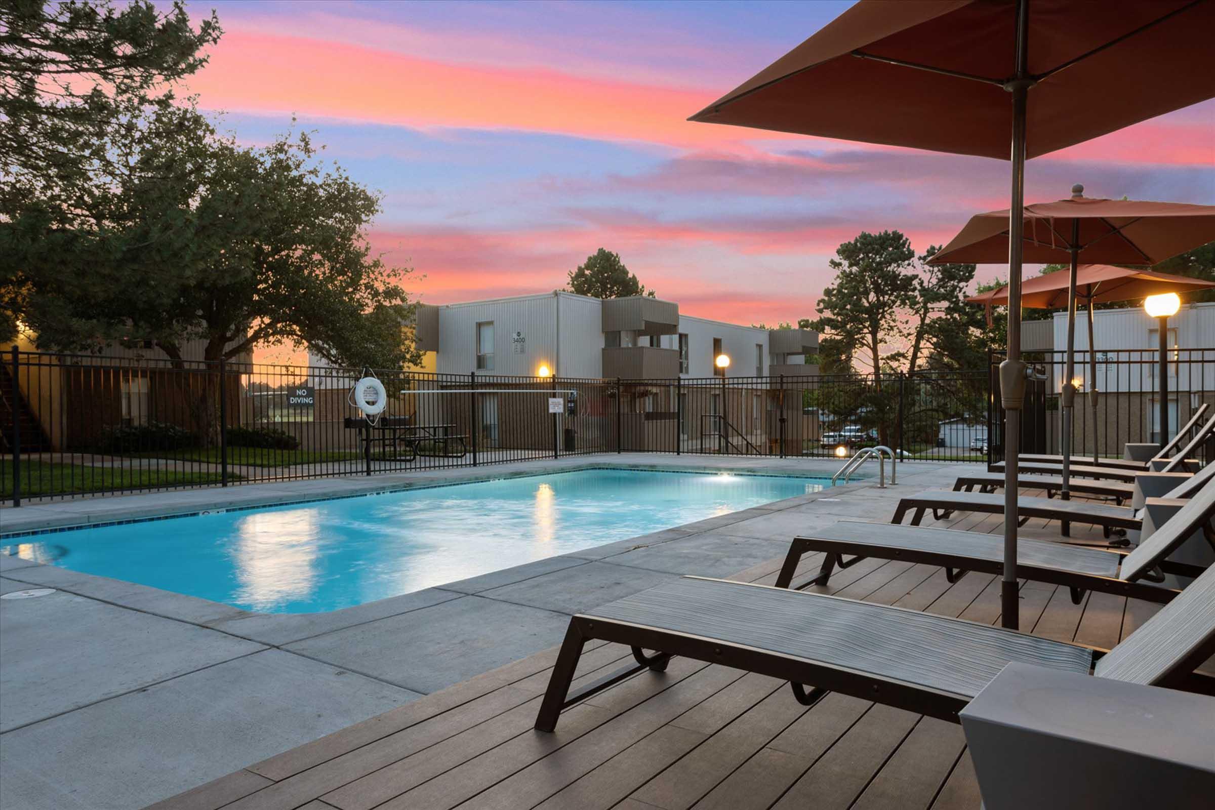 A serene pool area during sunset, featuring a clear blue pool surrounded by wooden lounge chairs and umbrellas. Lush green trees and apartment buildings are visible in the background, enhancing the tranquil outdoor setting. The sky displays vibrant pink and purple hues, creating a peaceful atmosphere.