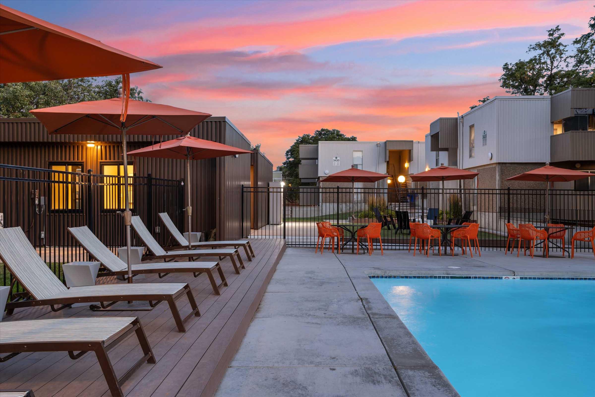A modern swimming pool area with lounge chairs and orange umbrellas against a colorful sunset sky. The pool is surrounded by a fence, and there are outdoor seating areas with orange chairs nearby. Stylish buildings are visible in the background, highlighting a relaxed atmosphere.