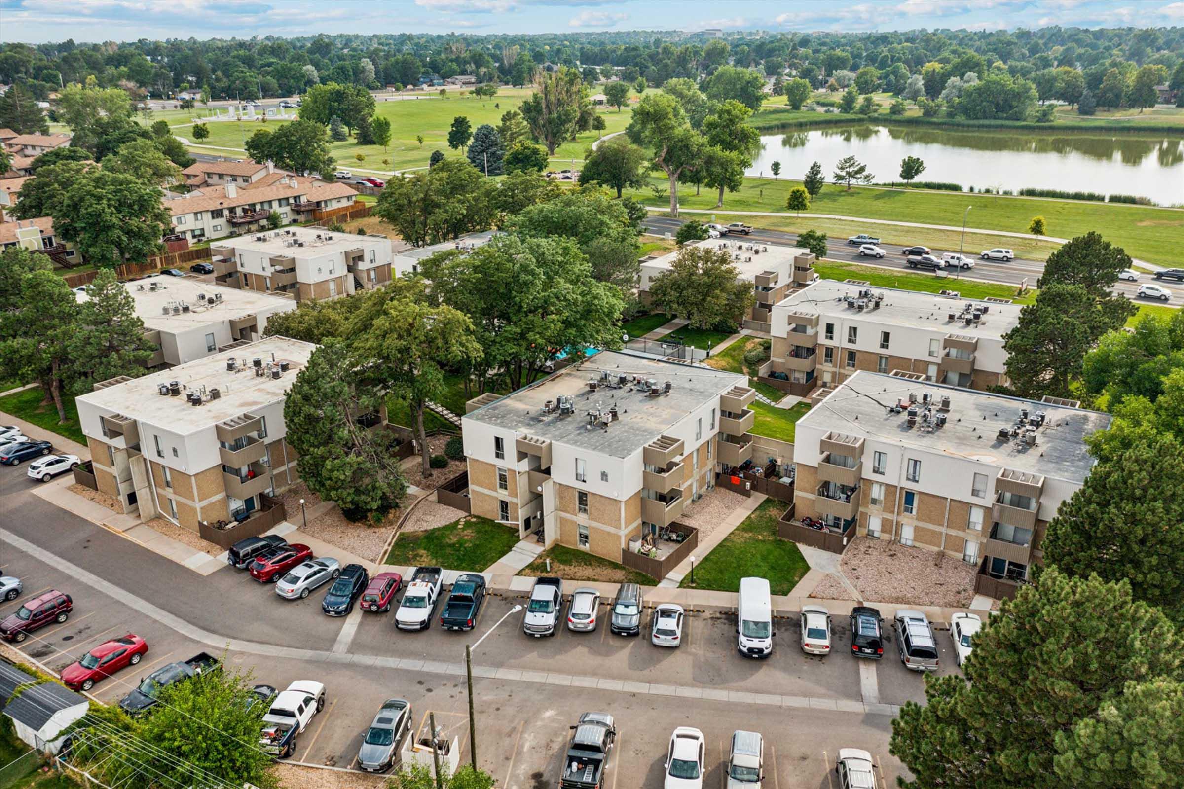 Aerial view of a residential area featuring multiple apartment buildings with flat roofs. Surrounding trees provide greenery, and there are parked cars along the street. In the background, a park area with a pond and expansive grassy fields can be seen. The scene captures a peaceful, suburban atmosphere.