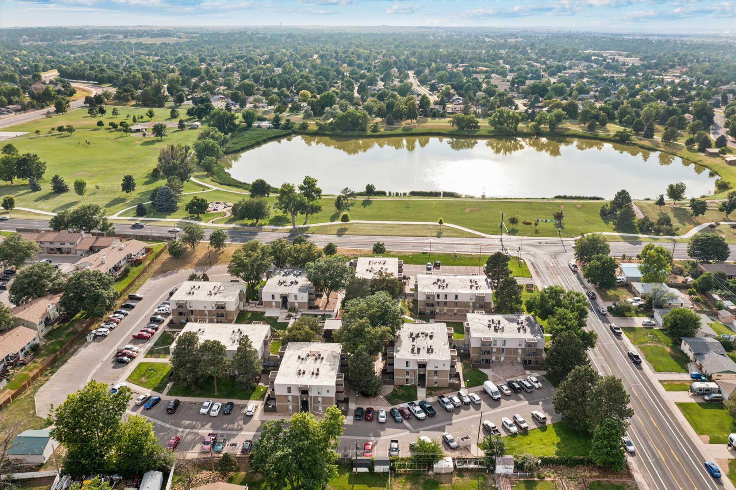 Aerial view of a residential area featuring several apartment buildings surrounded by trees and parking lots. In the background, a large pond reflects the sky, and a green park area is visible. The scene captures a suburban landscape with houses and roads extending into the distance.