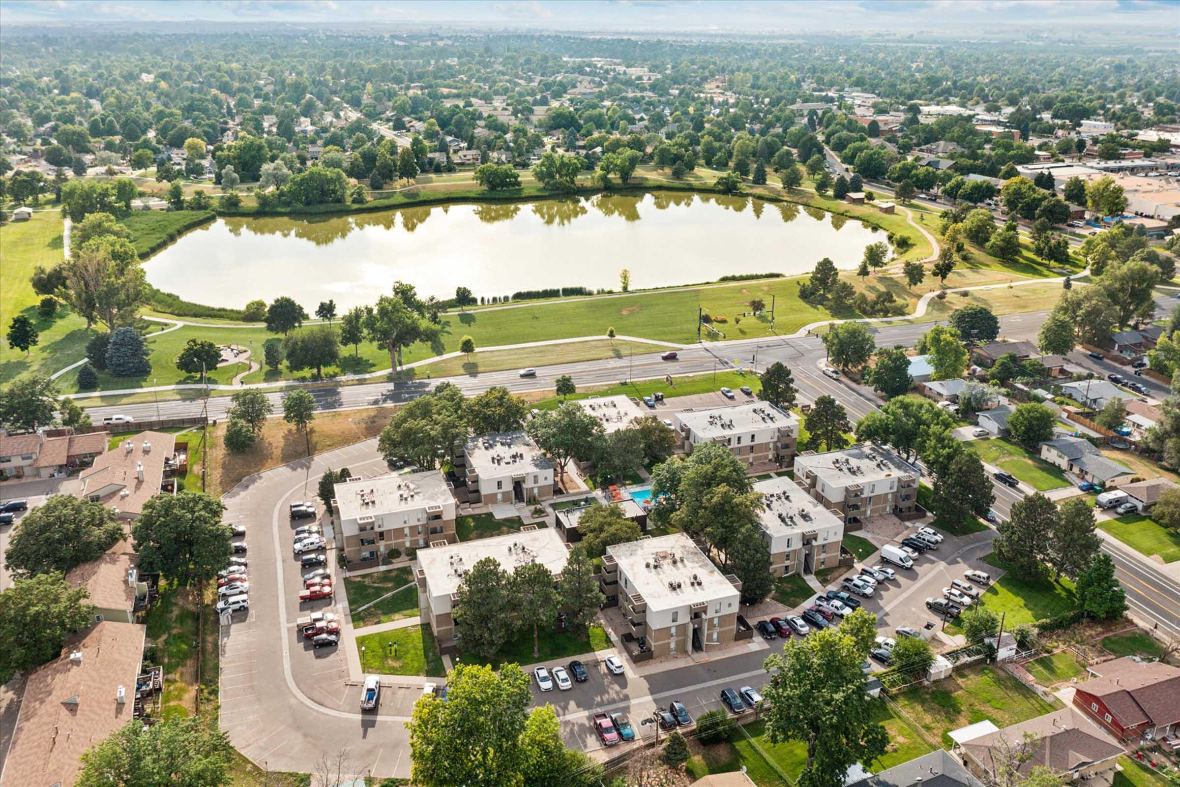 Aerial view of a residential area featuring multiple apartment buildings with parking lots, surrounded by trees. A large, serene pond is visible in the background, along with grassy parks and a road lined with houses. The landscape shows a mix of urban and green spaces under a partly cloudy sky.