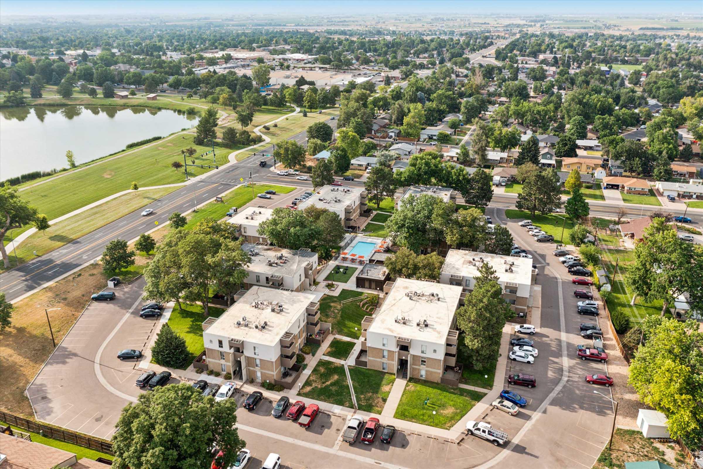 Aerial view of a residential area with multiple apartment buildings surrounding a swimming pool. Green spaces and trees are visible, alongside a road and a nearby lake. Vehicles are parked in a lot, and the landscape extends into the distance, showcasing urban and natural features.