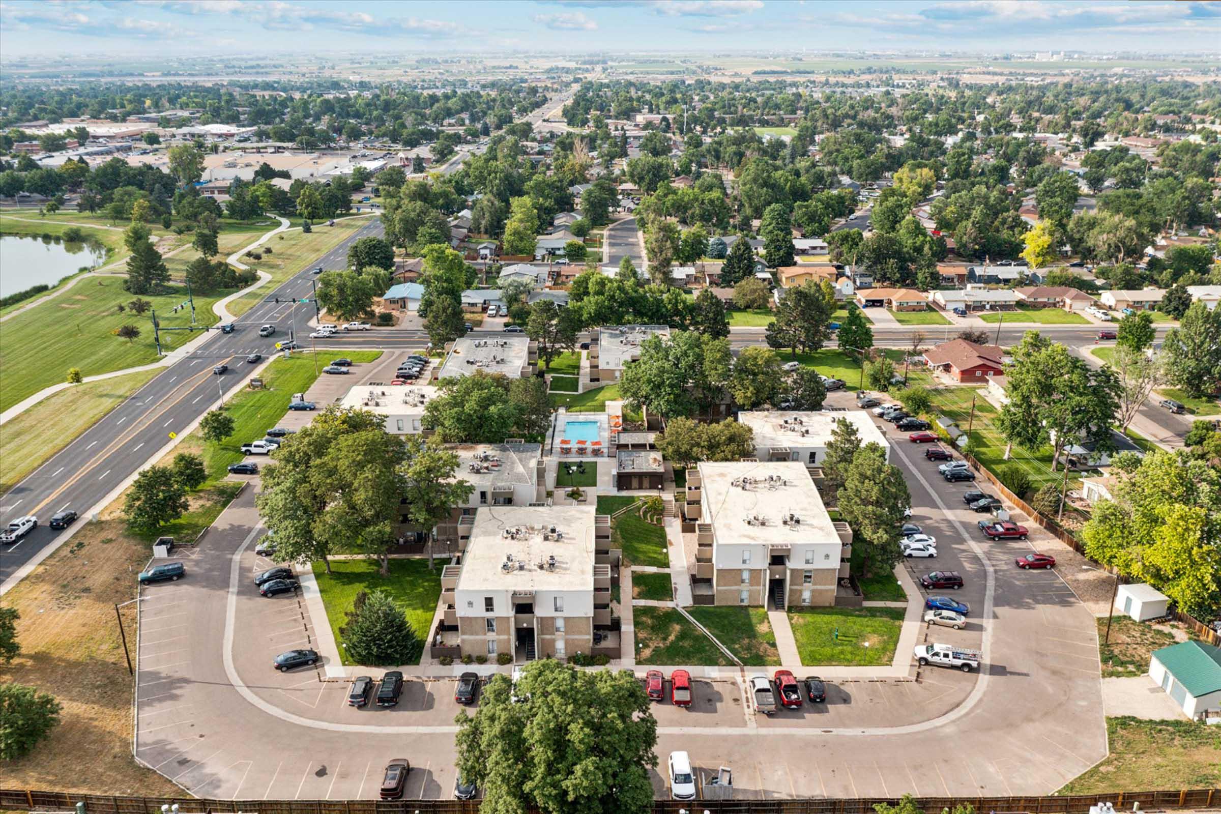 Aerial view of a residential apartment complex with multiple buildings arranged around a central courtyard, featuring green lawns and a swimming pool. Surrounding areas include roads, parked cars, and residential homes, set against a backdrop of trees and a distant landscape.