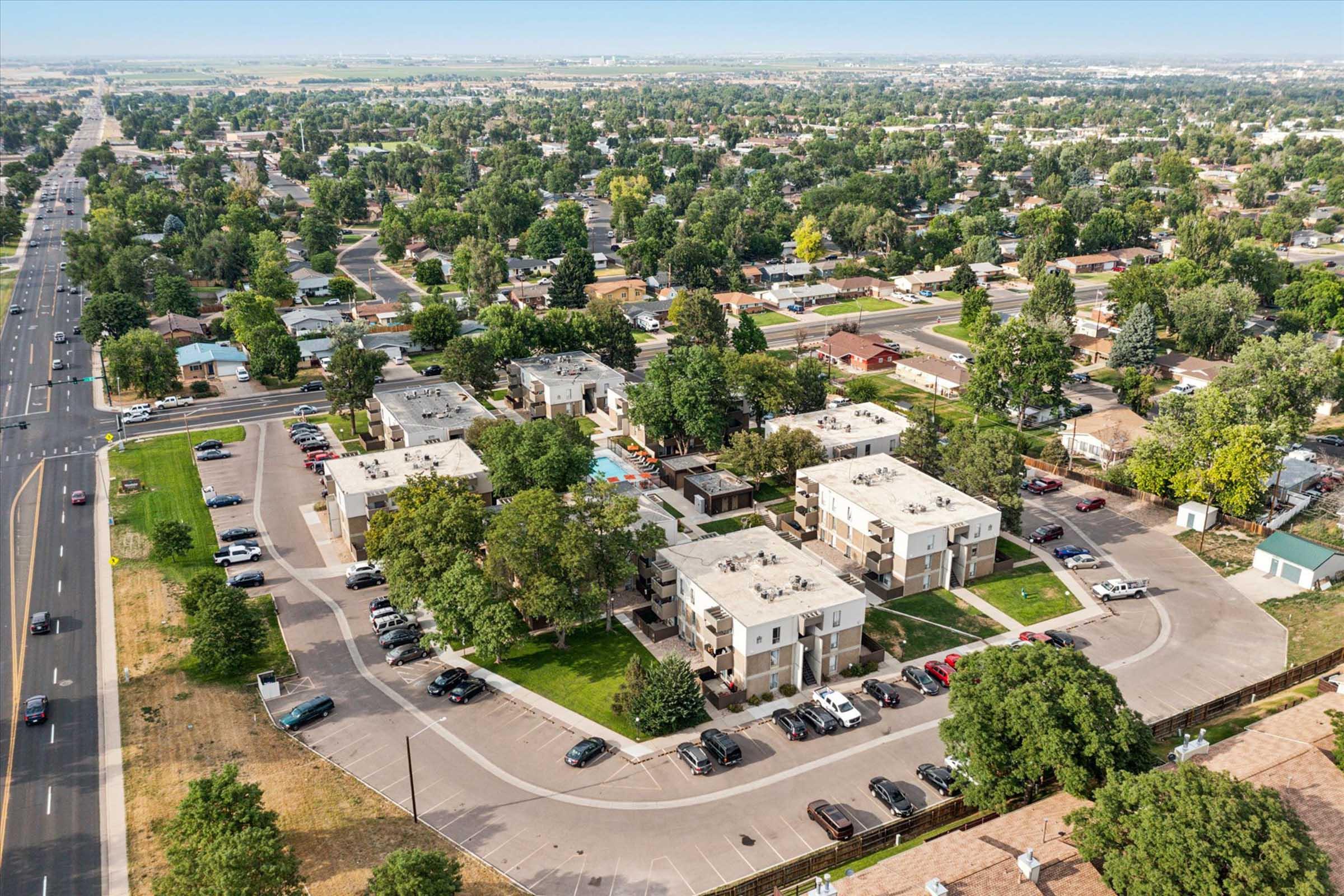 Aerial view of a residential area featuring multiple apartment buildings surrounded by trees and landscaped grounds. Roads and parked cars are visible, with a swimming pool area in the complex. In the background, additional homes and greenery stretch out, indicating a suburban neighborhood.