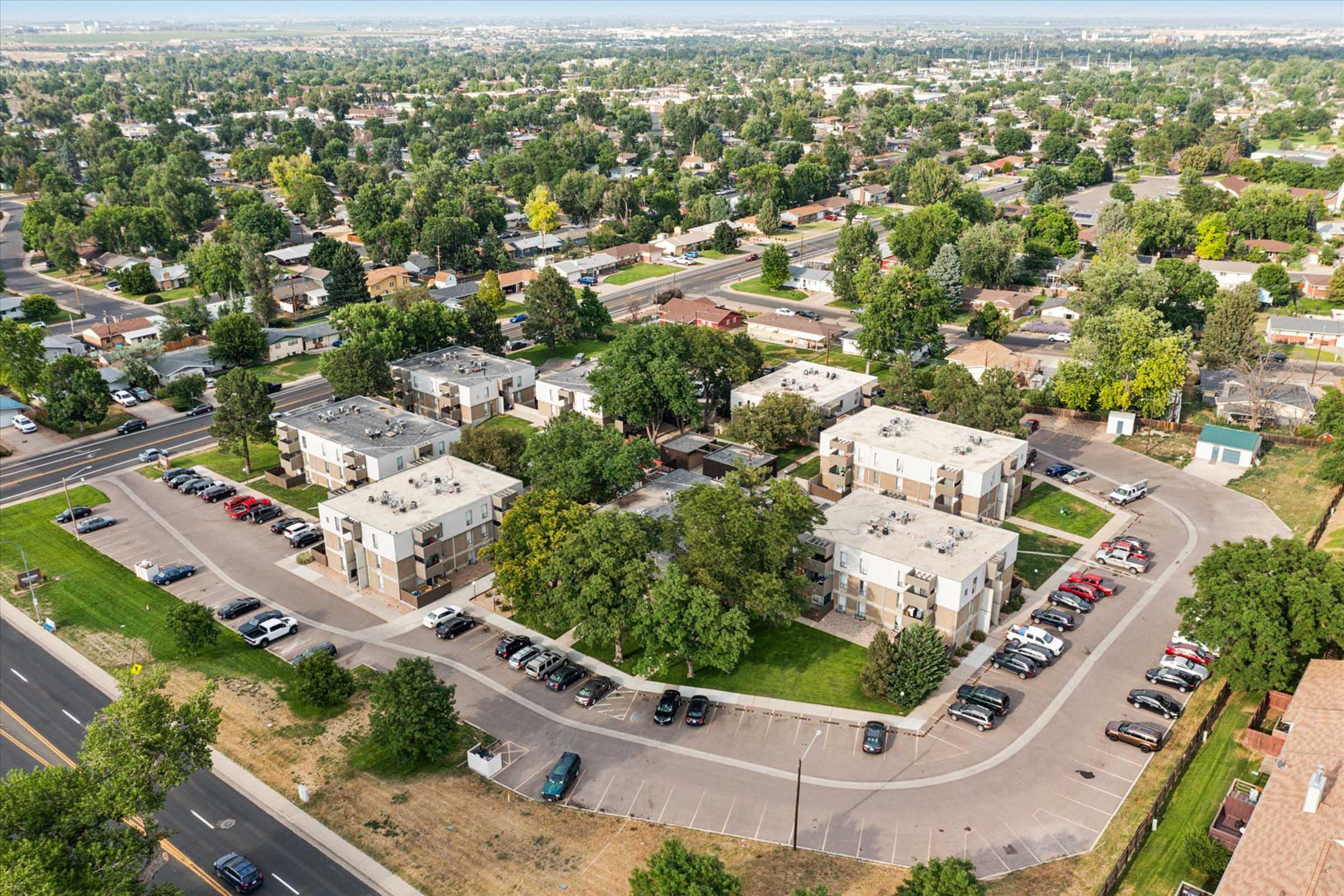 Aerial view of a residential complex surrounded by green trees and parked cars. The area features multiple buildings, with a mix of asphalt and grassy areas, set in a suburban environment with other houses and roads visible in the background.