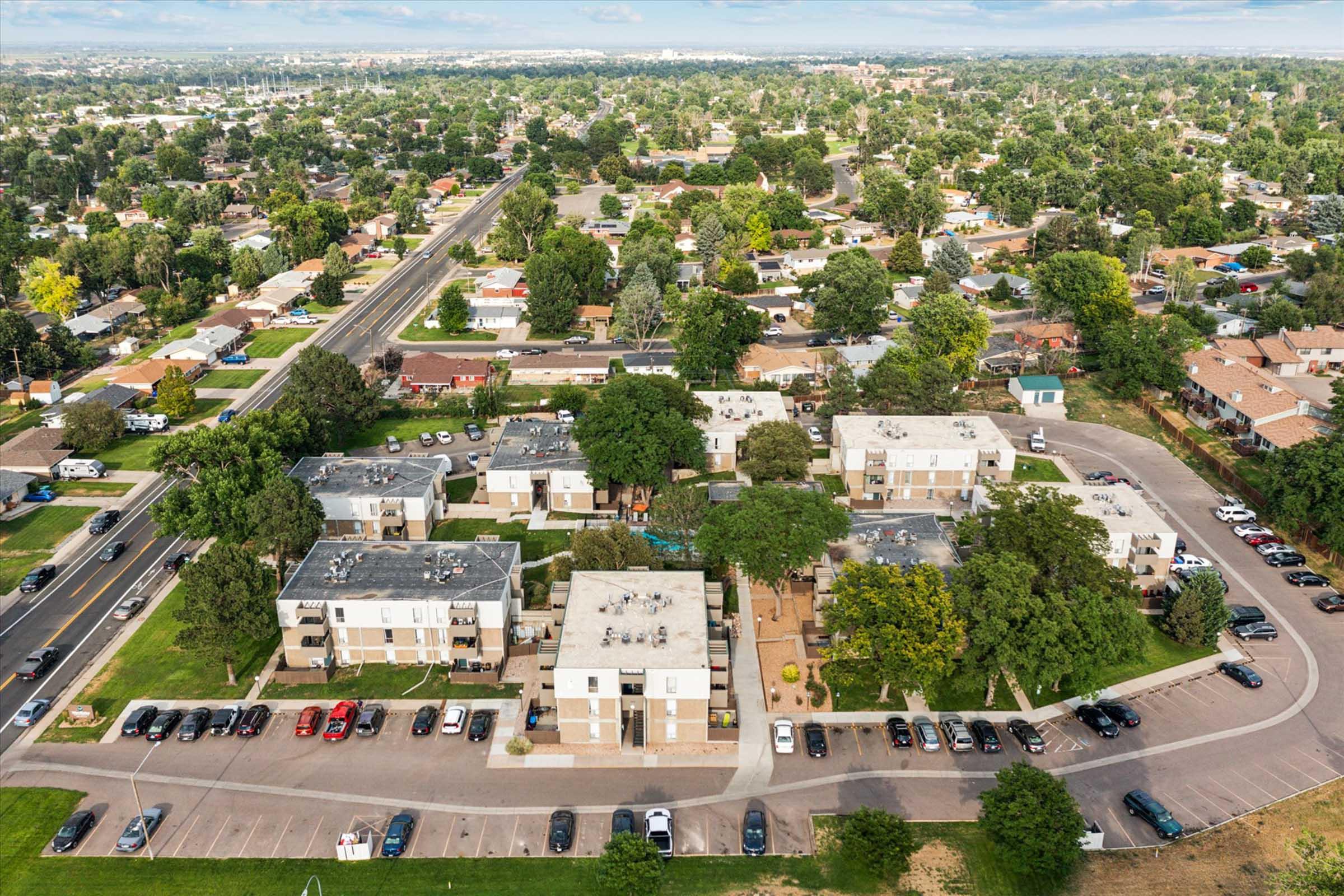 Aerial view of a neighborhood featuring residential apartment buildings surrounded by greenery. The area includes various trees, parked vehicles, and nearby streets. In the background, suburban homes and additional greenery can be seen, showcasing a typical residential community.