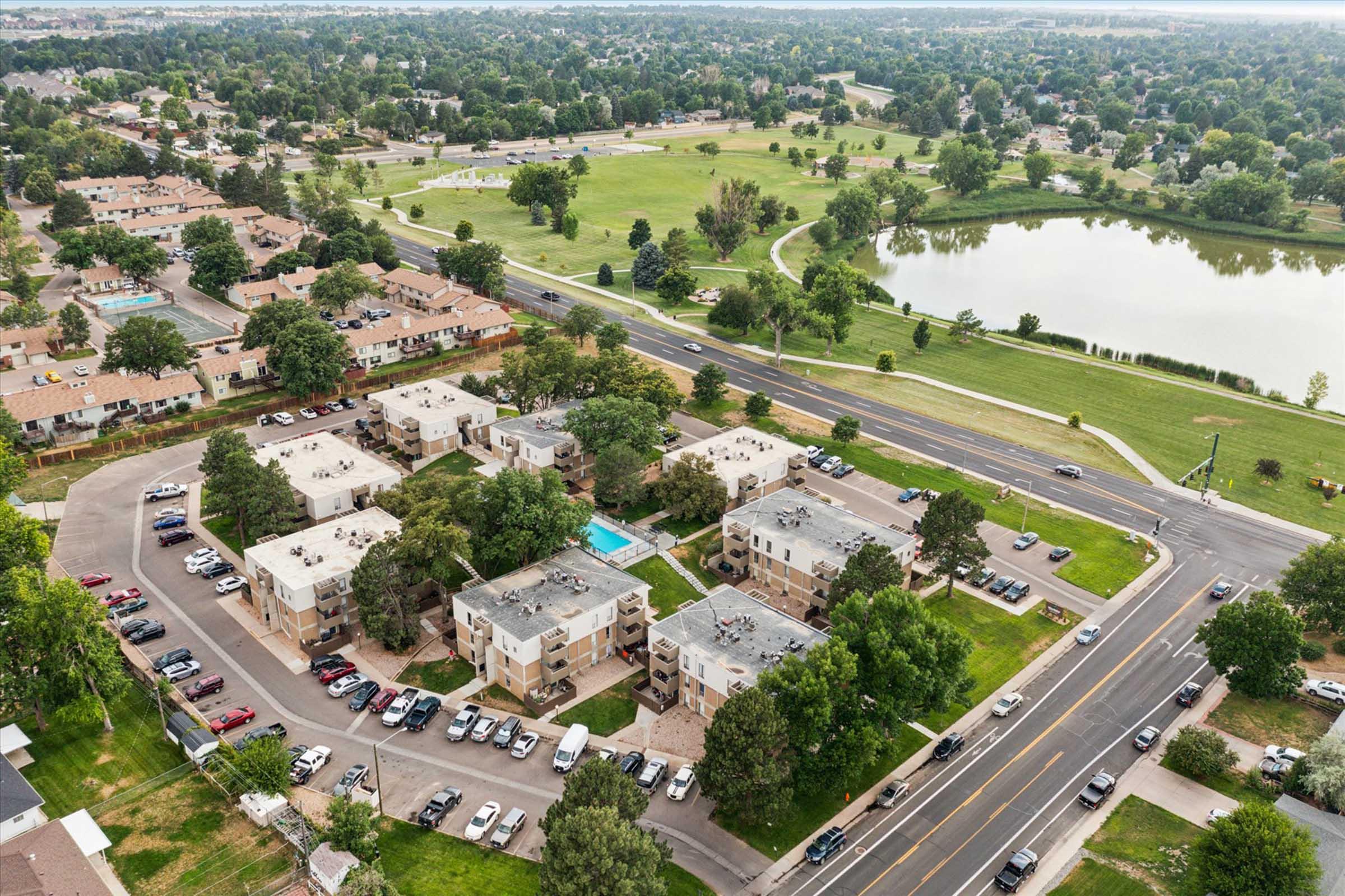 Aerial view of an apartment complex near a park and a pond. The complex features several buildings with green lawns and trees surrounding them. A road runs alongside the complex, with parked cars visible. The park includes walking paths and open green spaces.