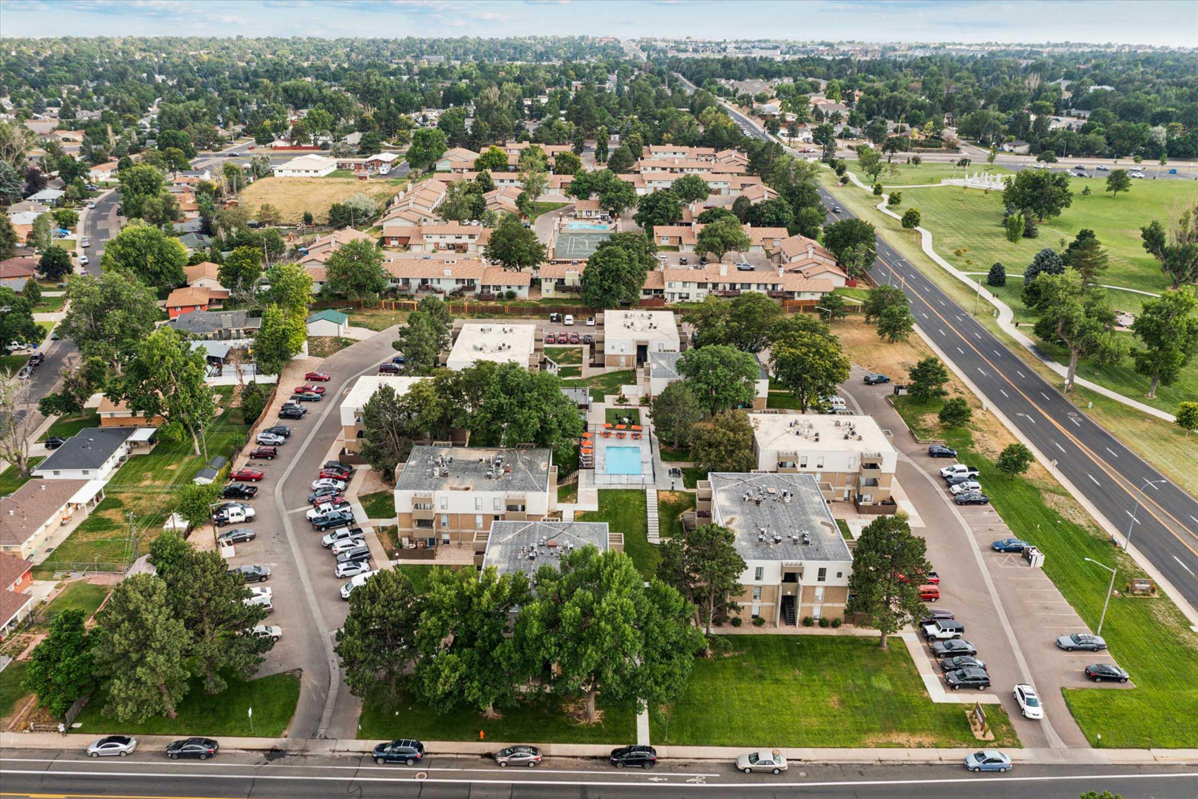 Aerial view of a residential area featuring several apartment buildings arranged around landscaped green spaces and a pool. Nearby streets are lined with parked cars, and the surrounding neighborhood includes additional homes and trees, with a golf course visible in the distance.