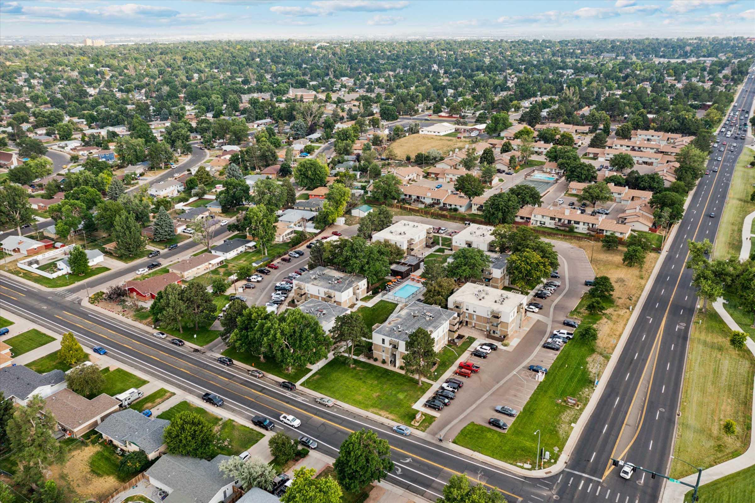 Aerial view of a suburban neighborhood featuring residential homes, green spaces, and roads. The scene includes well-maintained lawns, parked cars, and a community pool area visible among the buildings, along with trees lining the streets, indicating a developed and peaceful community environment.