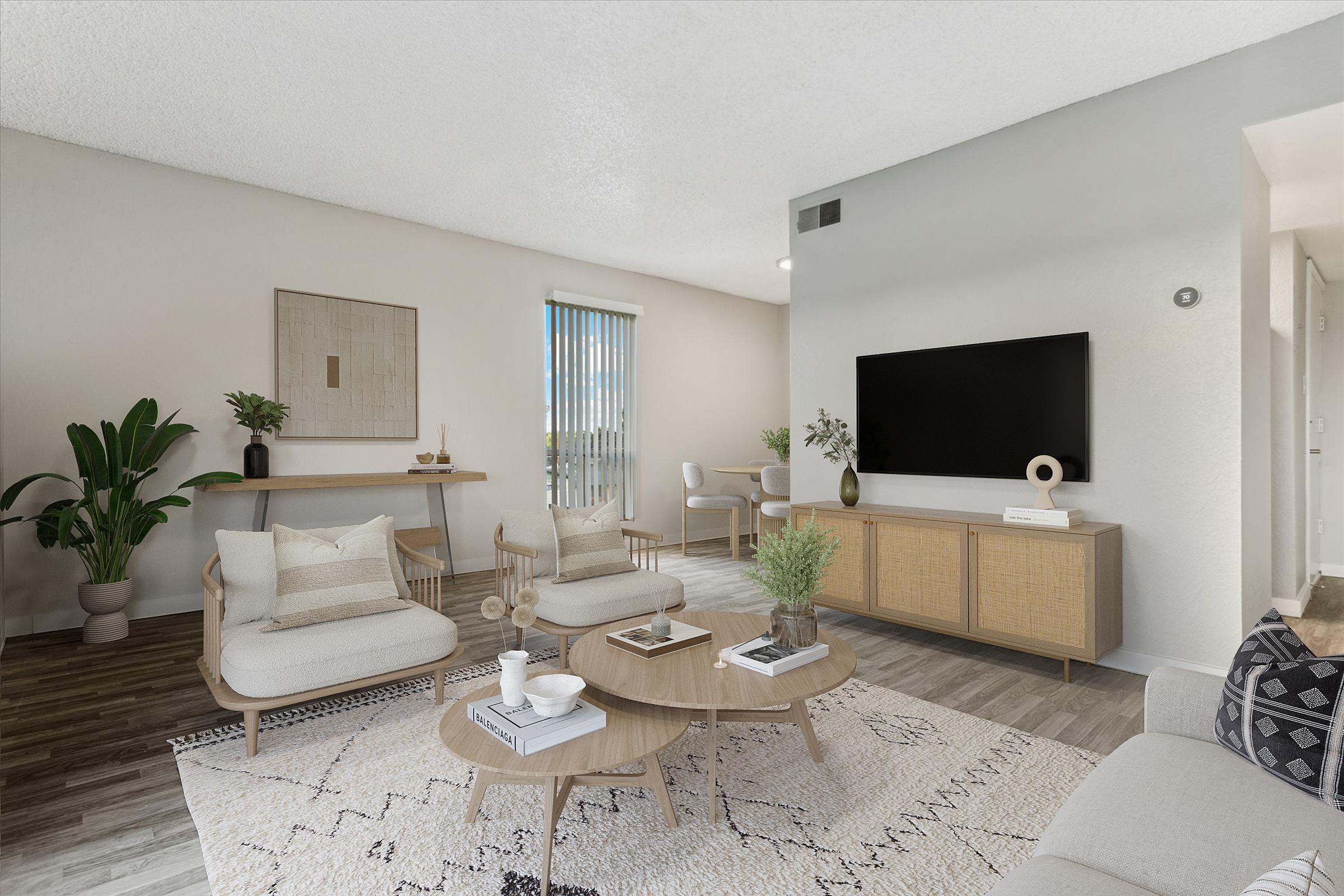 A modern living room featuring light-colored furniture, including two stylish chairs and a coffee table. There’s a large TV on a wooden console, decorative plants, and a patterned rug. The room is bright with natural light coming through a window adorned with vertical blinds. Minimalist decor enhances the inviting atmosphere.
