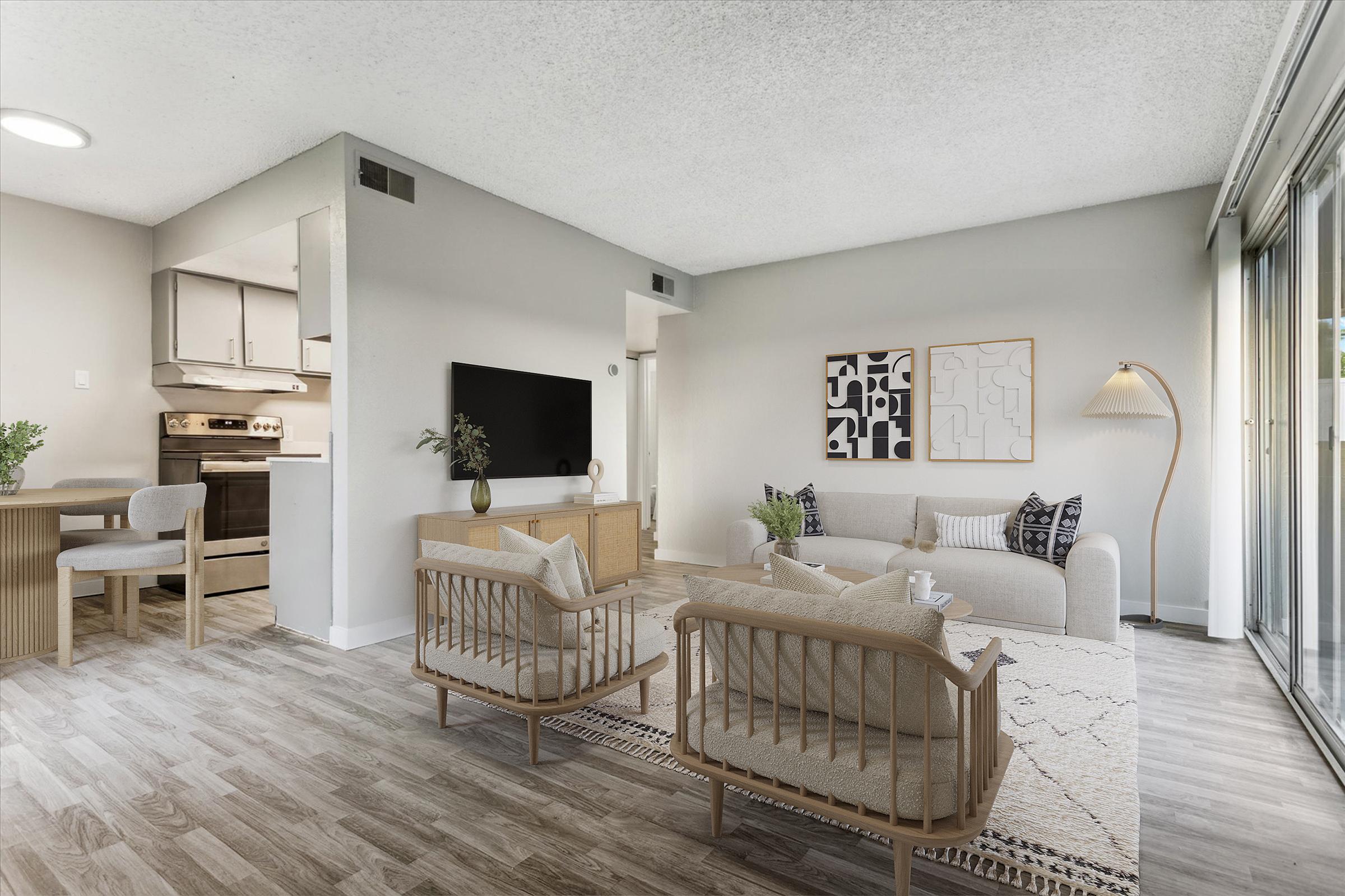 A modern living room with light-colored walls and wooden flooring. It features a comfortable sofa, two armchairs, a coffee table, and a large TV on a cabinet. A dining area is visible in the background with a simple table and chairs. Large windows let in natural light, and decorative plants enhance the space.