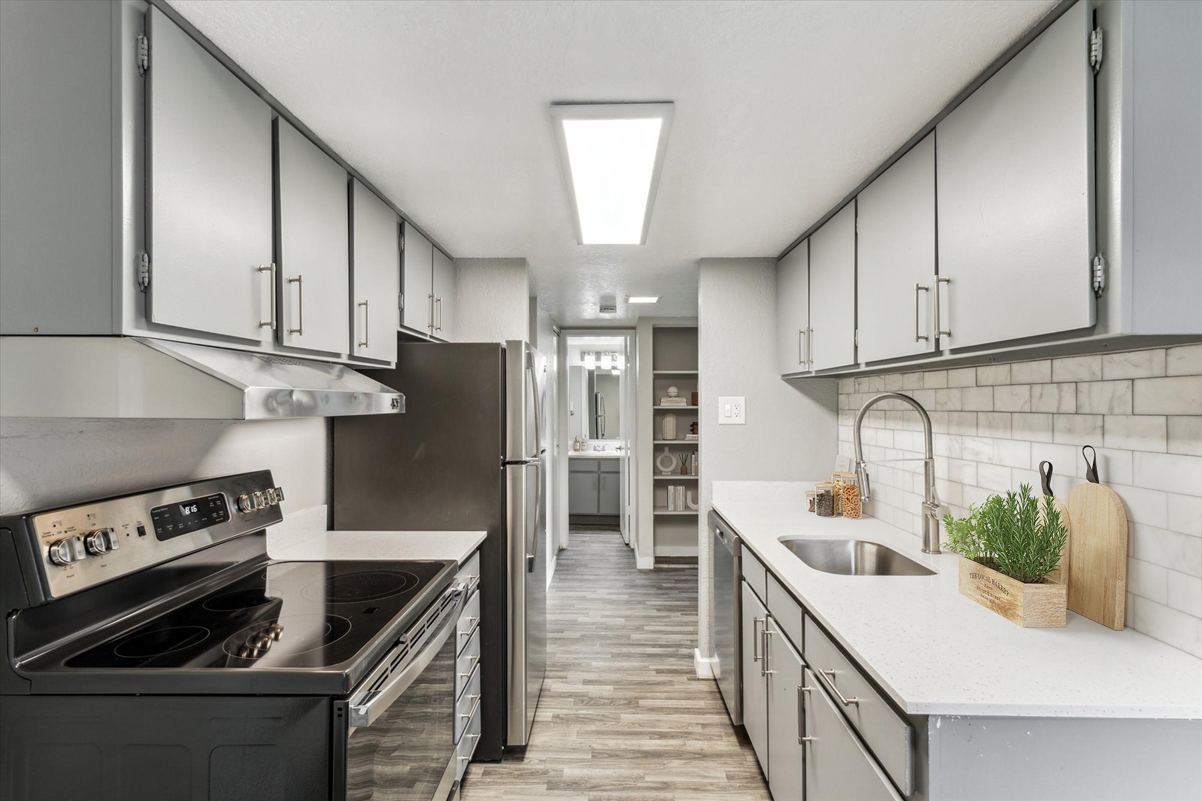A modern kitchen featuring grey cabinets, stainless steel appliances, and a marble-tiled backsplash. The space includes an electric stove, a double sink with a minimalist faucet, and a countertop with a small plant in a wooden pot. Ample natural light floods the area, enhancing its sleek design.