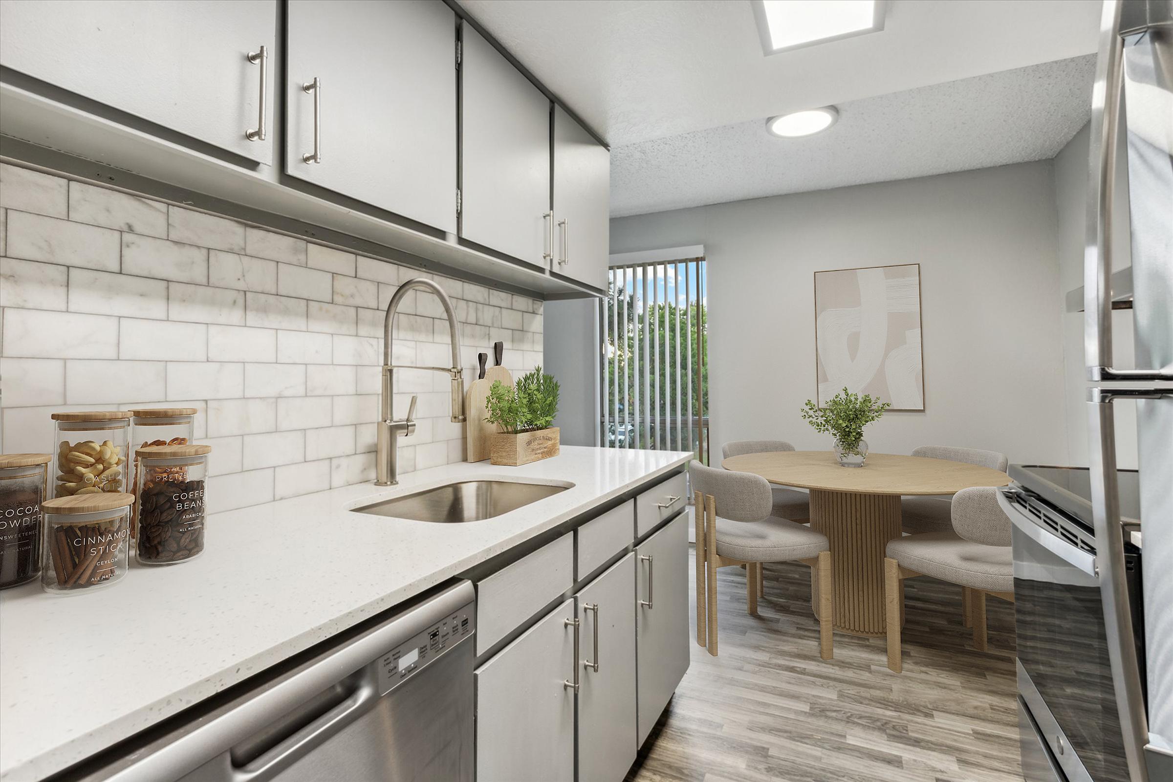 A modern kitchen featuring a white countertop with a stainless steel sink, gray cabinets, and a tiled backsplash. Glass jars containing snacks are placed on the counter. A small round dining table with four upholstered chairs is set nearby, complemented by a plant and artwork on the wall. Natural light enters through a window.