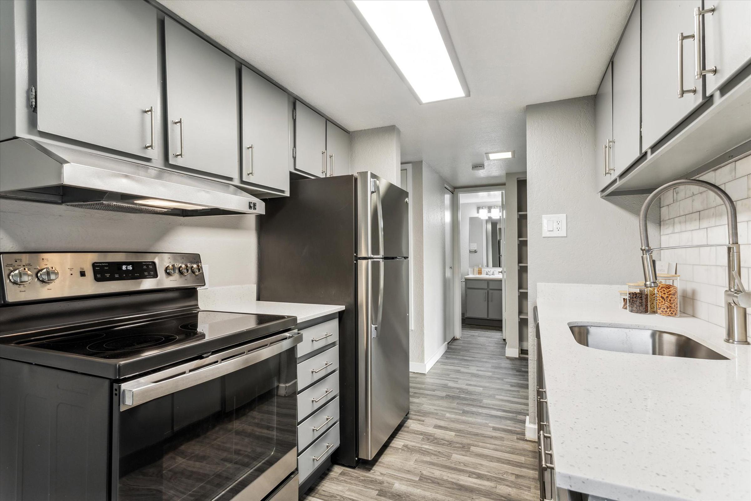 Modern kitchen featuring gray cabinetry, stainless steel appliances including an oven and refrigerator, a white countertop, and a sleek undermount sink. The space is well-lit with overhead lighting, and a tiled backsplash adds a clean aesthetic. A hallway leads to another room in the background.