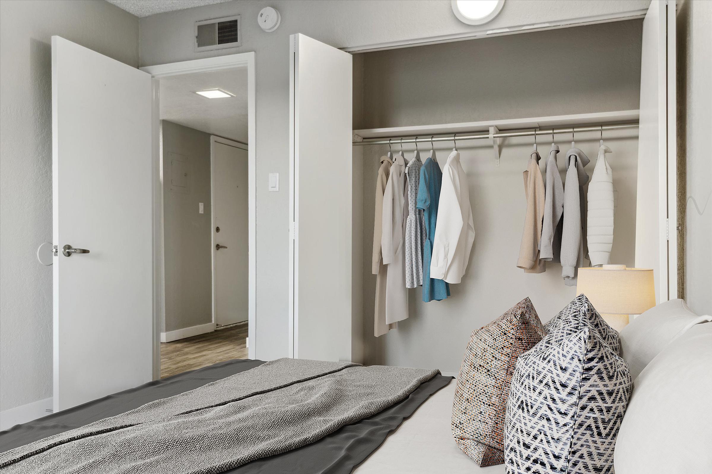 A cozy bedroom featuring a neatly made bed with decorative pillows, a gray bedspread, and a lamp on a bedside table. A closet with hanging clothes is visible, along with an open door leading to a hallway. The walls are painted in neutral tones, creating a calm atmosphere.