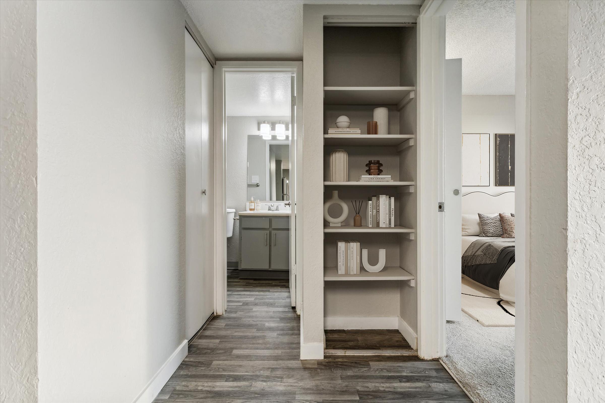 A view of a hallway in a modern apartment, featuring a built-in bookshelf with decorative items and books. On the left, a bathroom door is visible, while the right side leads to a cozy bedroom with a bed and soft decor. The flooring is a dark laminate, adding warmth to the space.