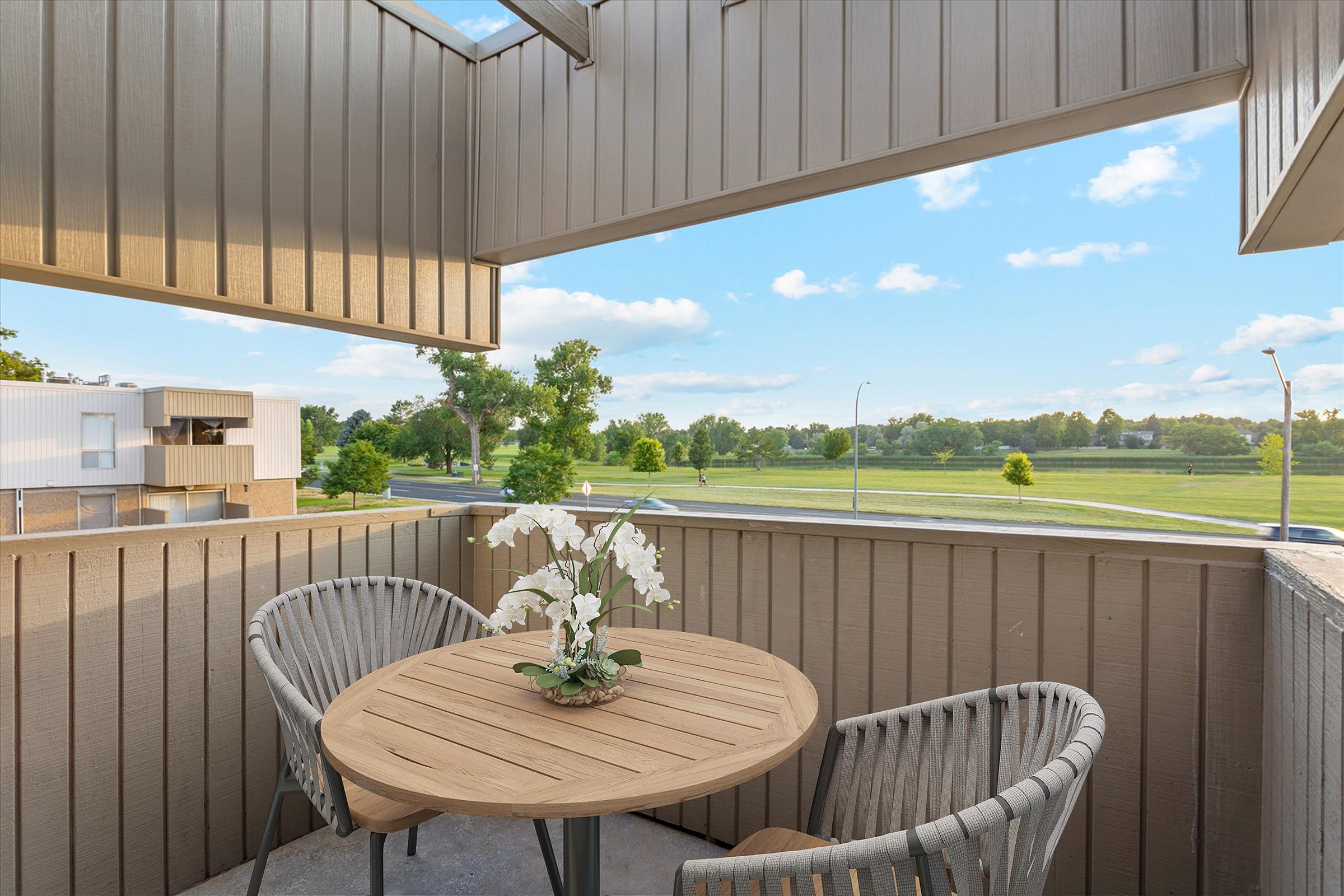 A cozy balcony view featuring a round wooden table with a small centerpiece of white flowers. Two stylish wicker chairs are arranged around the table. In the background, a green park and trees are visible under a blue sky with fluffy clouds, providing a serene outdoor setting.
