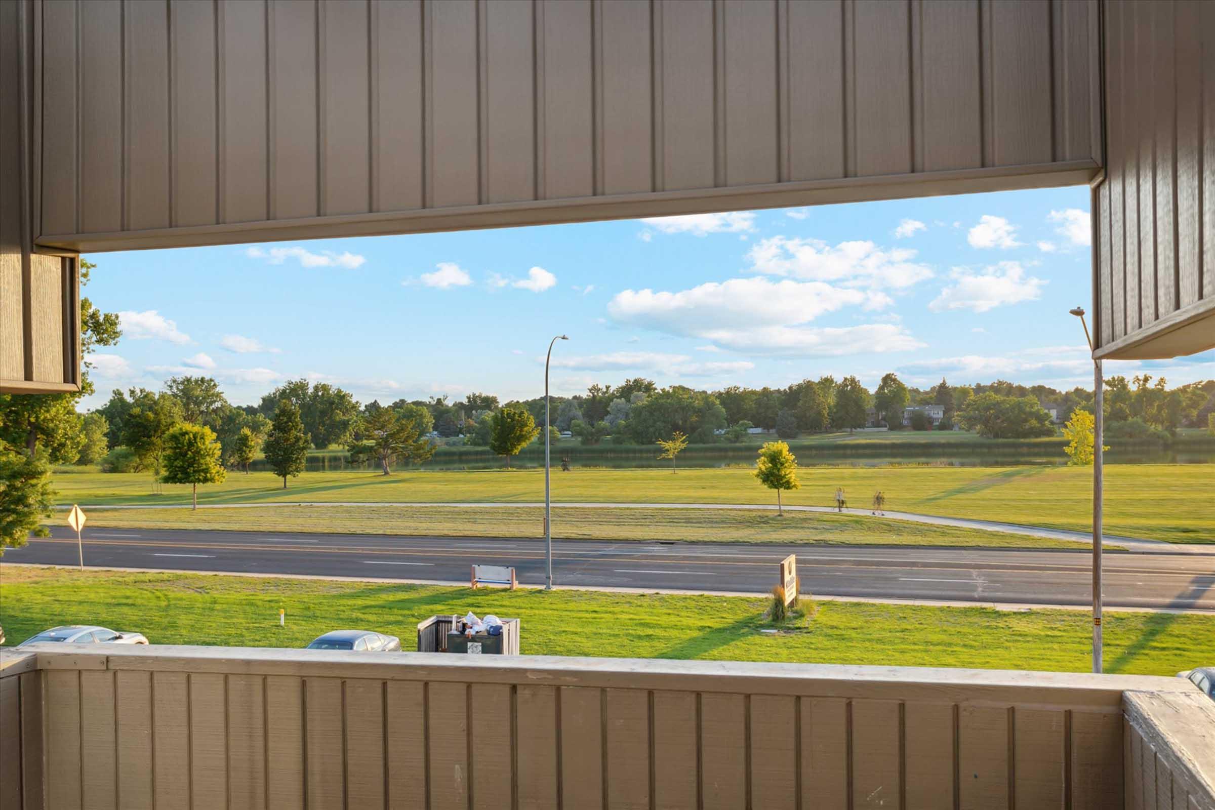 View from a balcony overlooking a grassy field with trees. A road runs parallel to the field, and blue skies with scattered clouds are visible in the background. The scene captures a tranquil outdoor setting, ideal for relaxation.