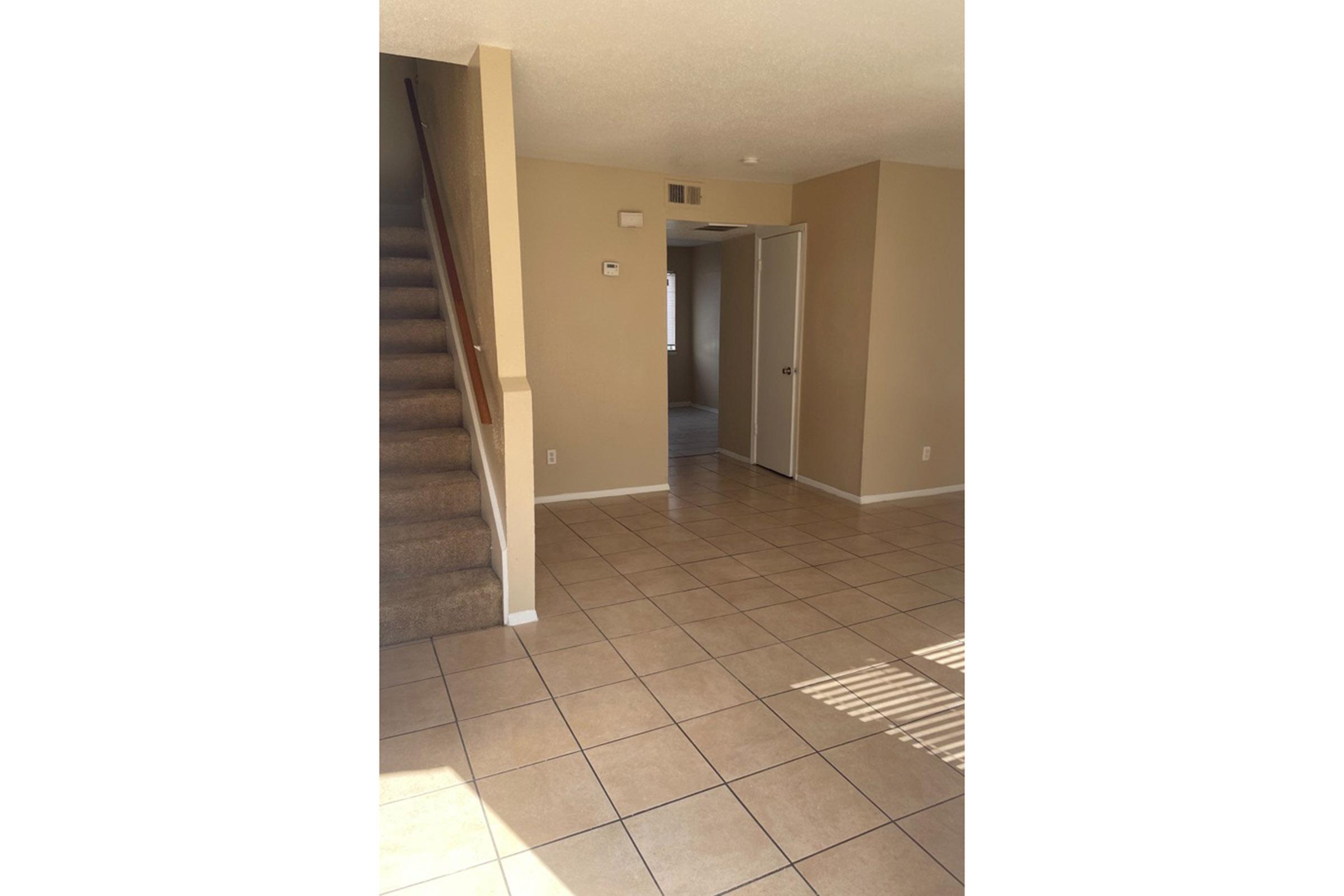 A view of an interior space featuring a staircase on the left, beige walls, and tiled flooring. The room has a doorway leading to another area, with natural light coming through a window. The overall atmosphere is bright and spacious, suitable for living or dining.