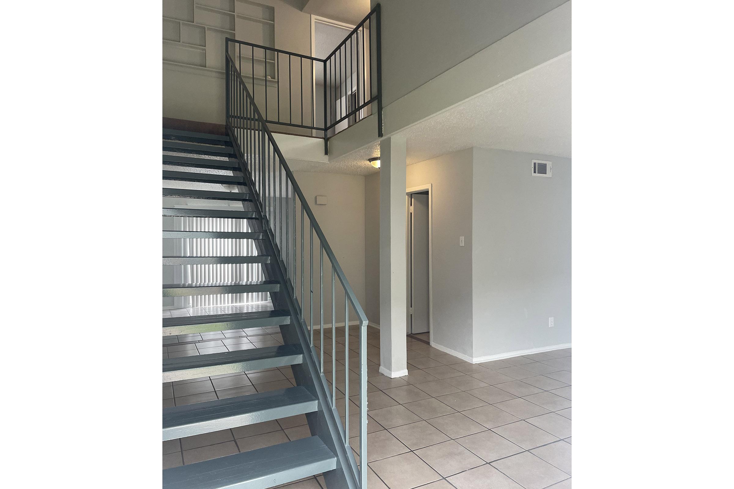 Interior view of a modern two-story space featuring a staircase with blue railings on the left, tiled floor, and light gray walls. The open layout includes a doorway to the right and a small landing area at the top of the stairs, providing a bright and airy atmosphere.