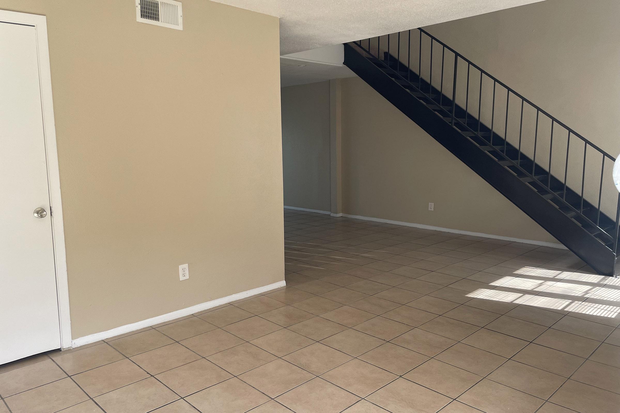A spacious interior of a home featuring beige walls and tiled flooring. There's a staircase on the right leading to an upper level, with natural light coming through a window. The room is empty with a clean and simple aesthetic.