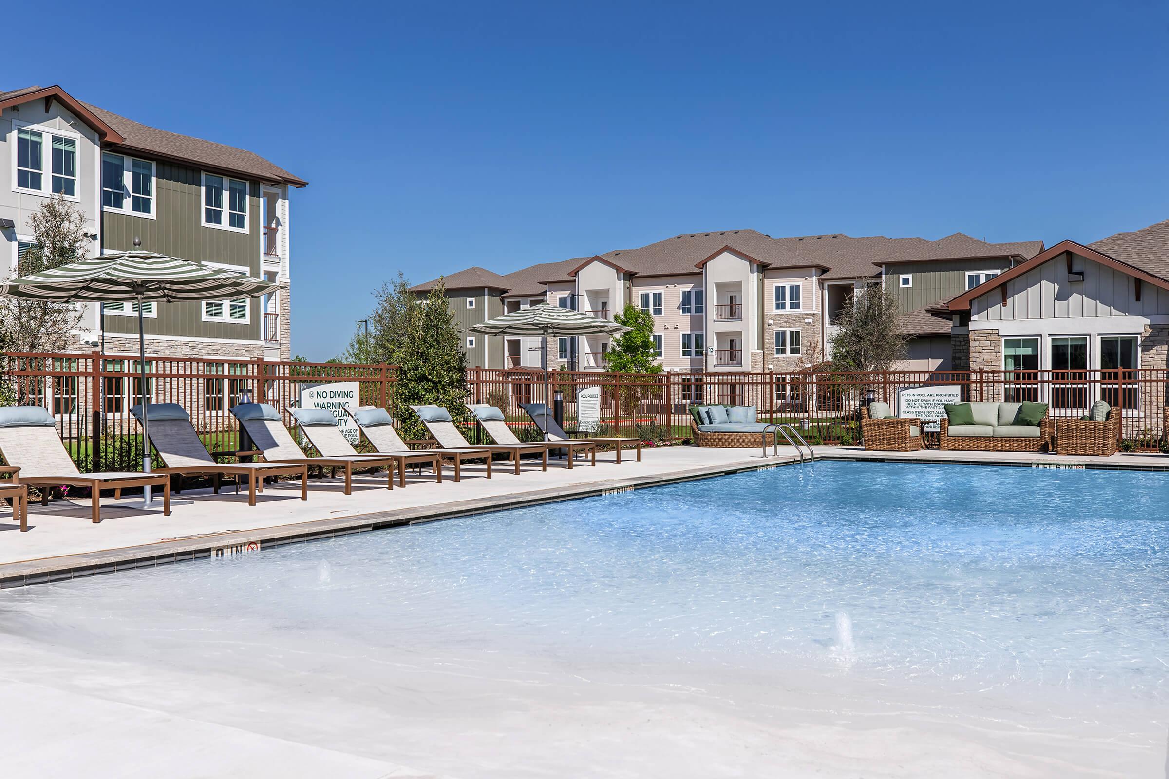 A sunny outdoor pool area surrounded by lounge chairs and umbrellas, with modern apartment buildings in the background. The pool has clear blue water and a welcoming sign, creating a relaxing atmosphere for residents and guests.