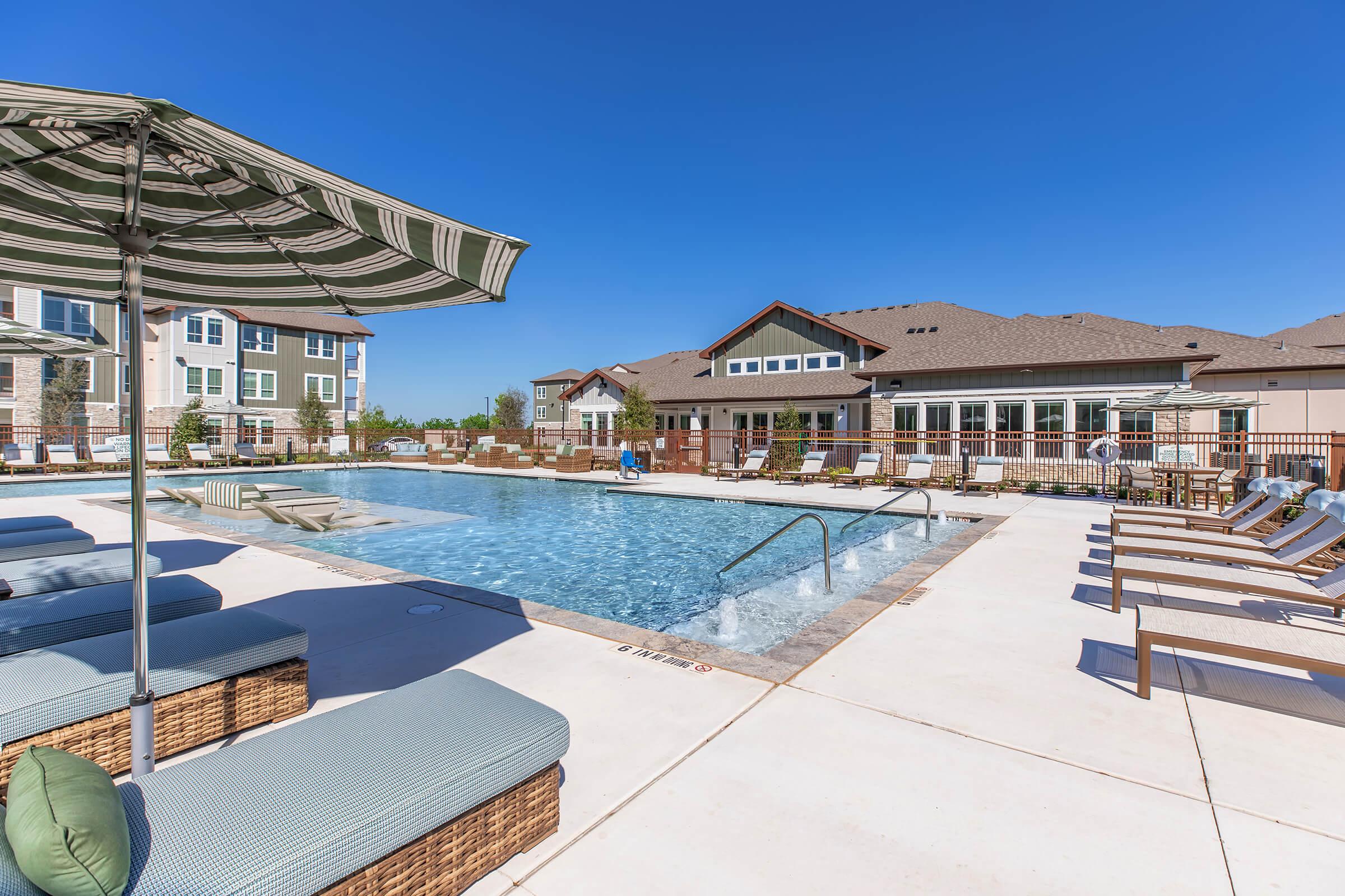 A sunny outdoor swimming pool area featuring lounge chairs and umbrellas. The pool is surrounded by a patio with a modern clubhouse in the background, showcasing a clear blue sky.