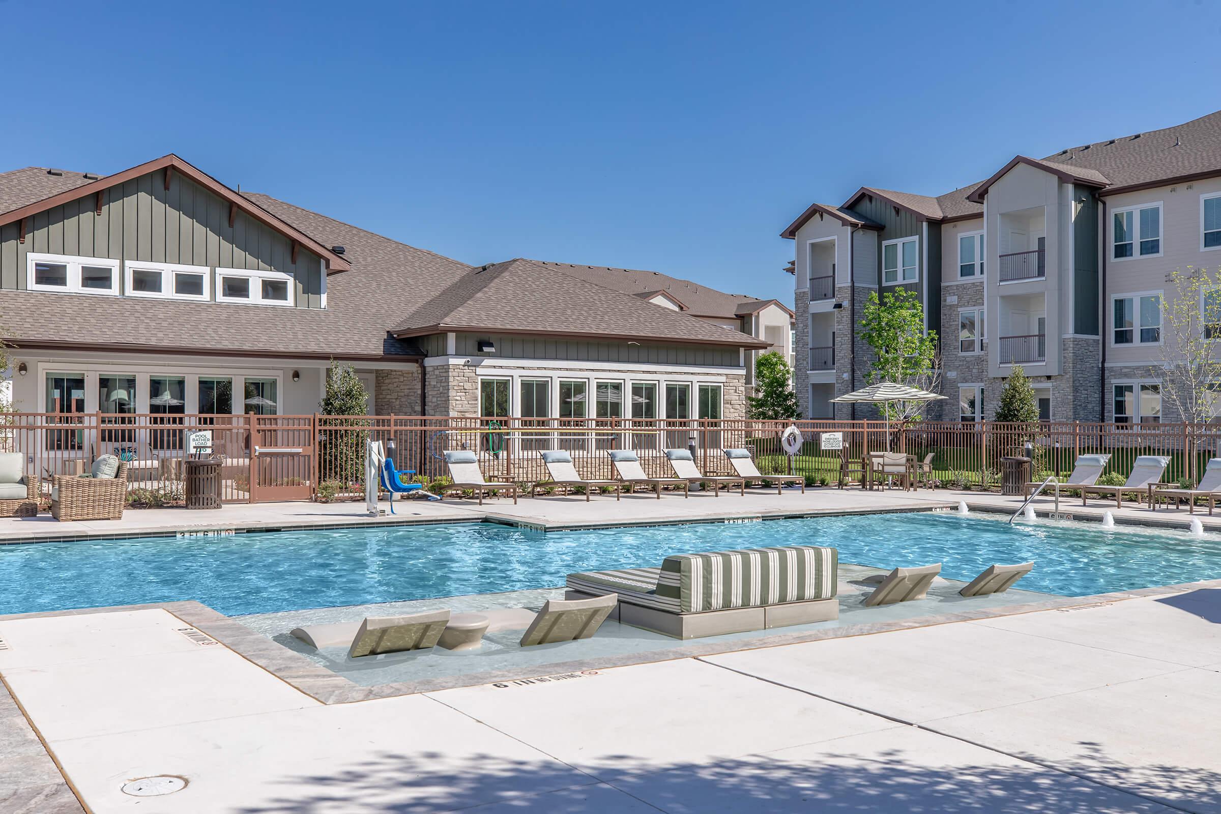 A clear blue swimming pool surrounded by lounge chairs and umbrellas, with a modern building in the background. The setting is sunny, featuring green grass and landscaping, creating a relaxing outdoor atmosphere. Ideal for leisure and social gatherings.