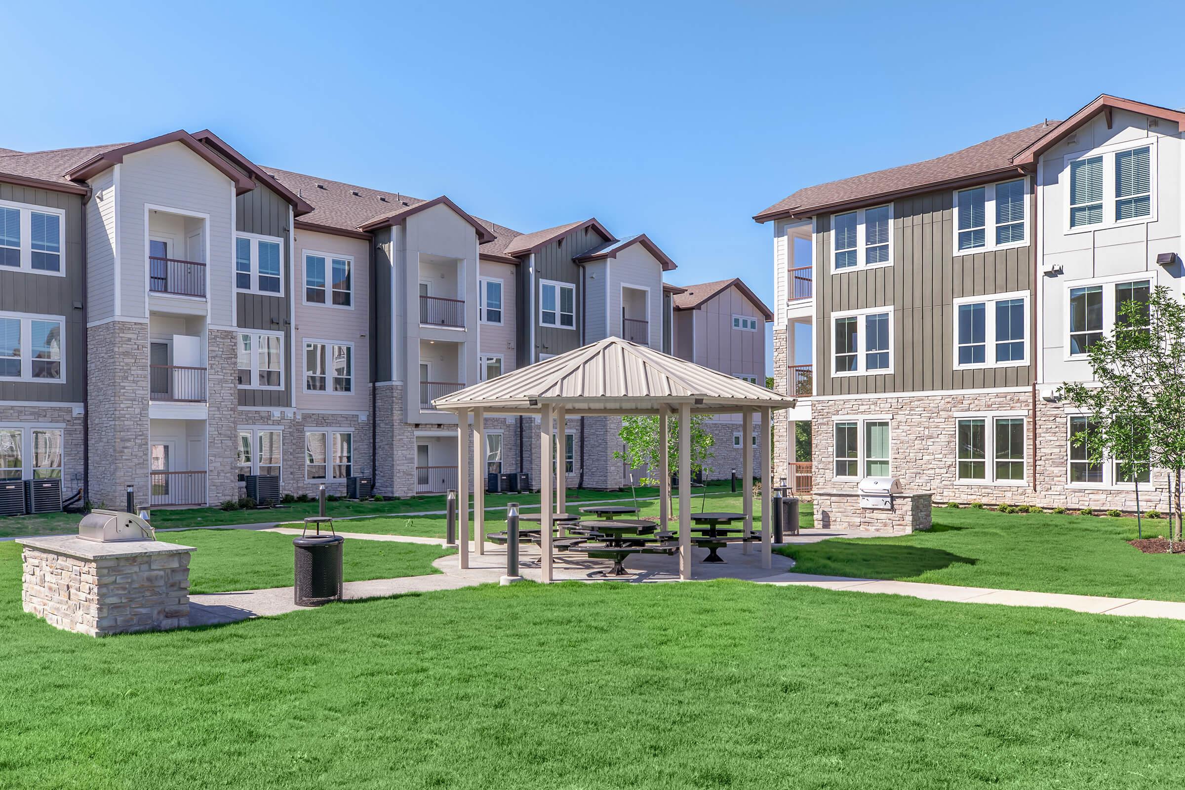 A landscaped area features a gazebo with picnic tables surrounded by green grass. In the background, modern apartment buildings showcase a mix of siding and stone exteriors under a bright blue sky. Trash bins and a grill are visible nearby, suggesting a communal outdoor space for residents.