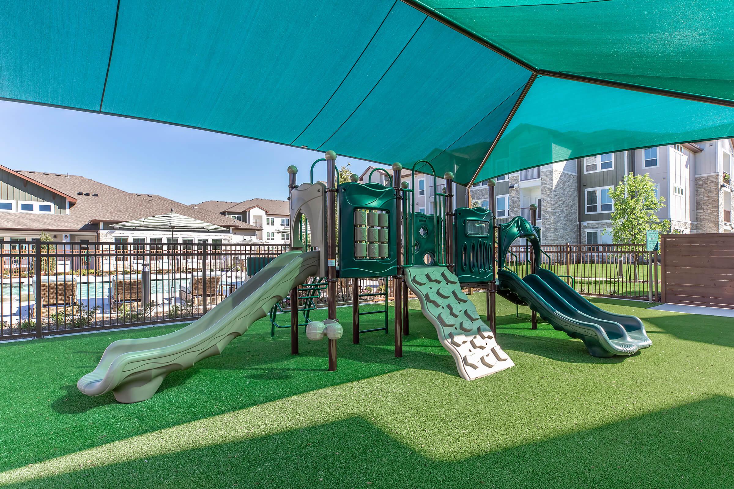 Playground featuring two green slides, climbing structures, and shaded with a large teal canopy. The area is surrounded by a safety fence and has artificial grass, with modern apartment buildings visible in the background. The scene is bright and inviting, perfect for children's play.