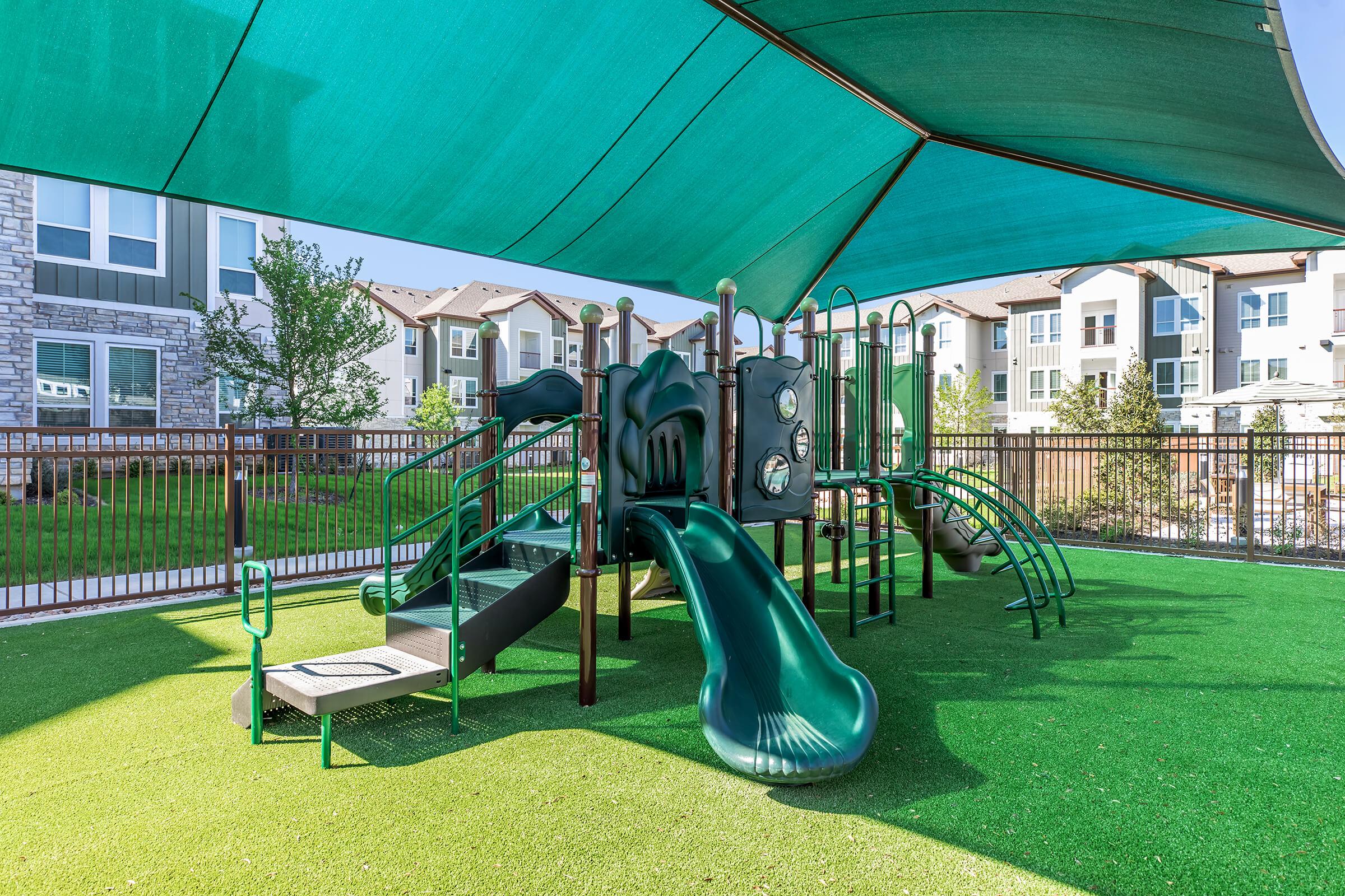 A colorful playground structure featuring slides, climbing features, and various play elements, set on a lawn with artificial grass. The area is shaded by a large canopy and is surrounded by a fence with residential buildings in the background.