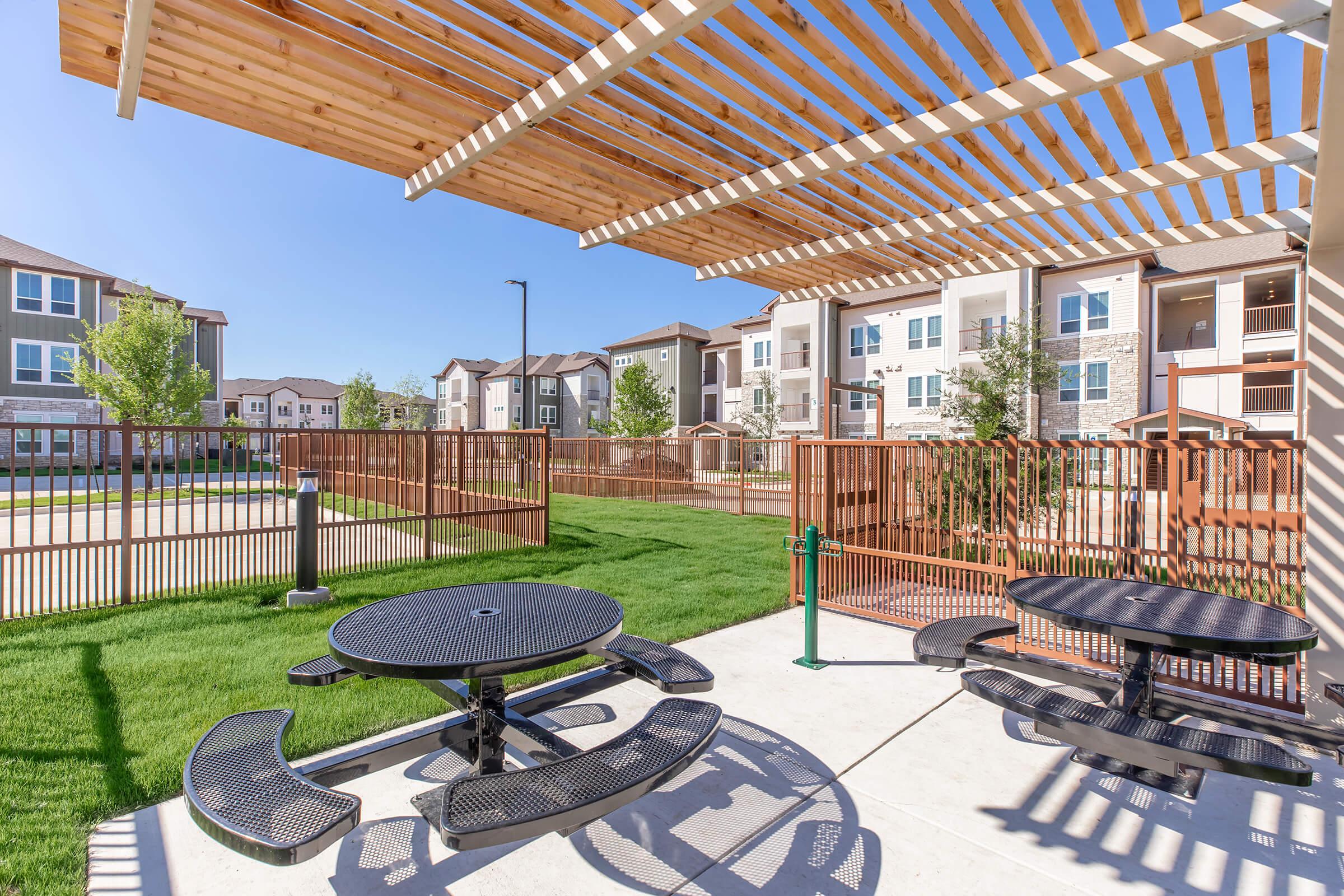 Outdoor seating area with black picnic tables under a wooden slatted pergola. Green grass and a fence surround the area, with a view of modern multi-unit apartment buildings in the background. Clear blue sky enhances the inviting atmosphere of the space.