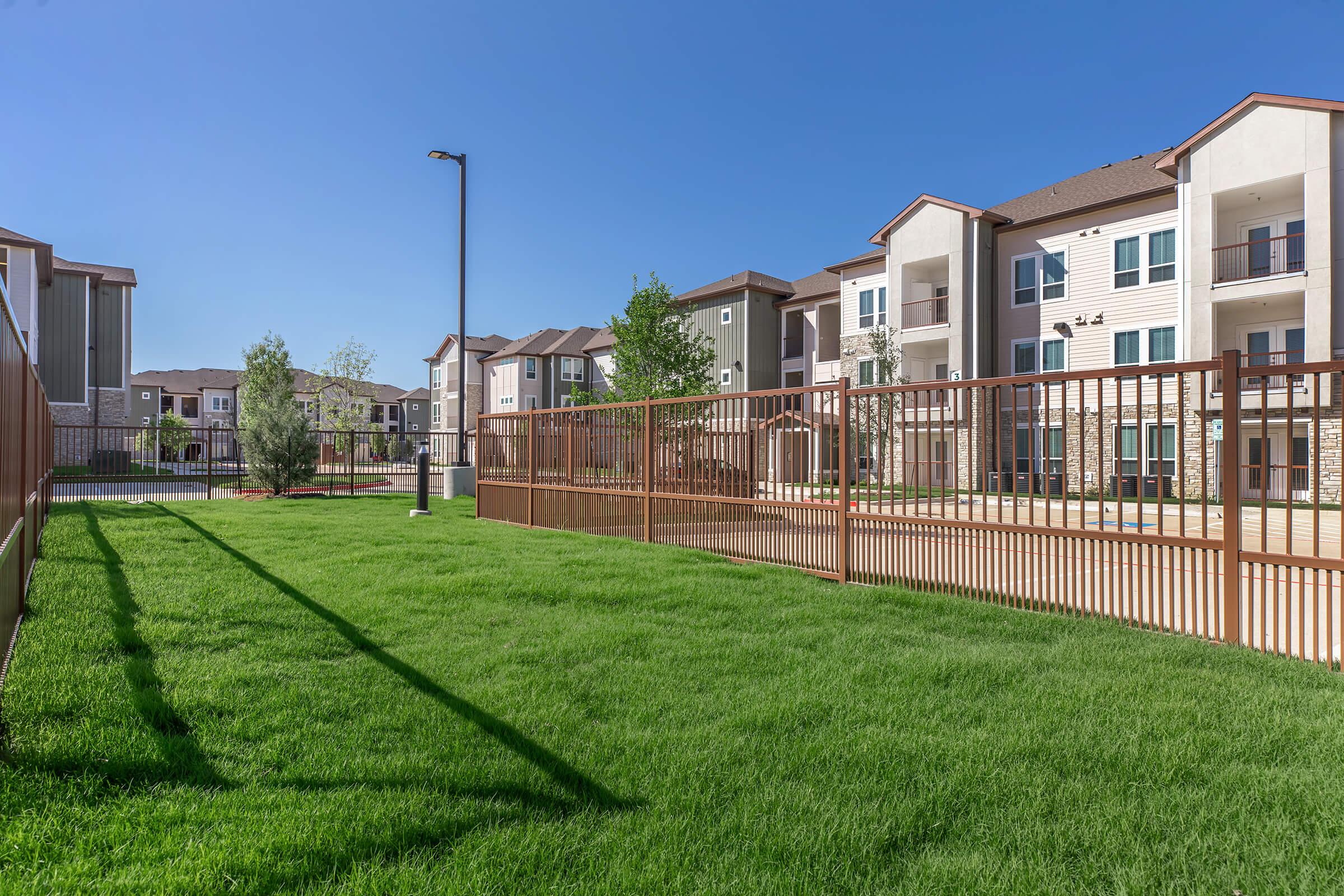 A well-maintained green lawn in front of a fenced area, leading up to modern apartment buildings under a clear blue sky. The apartments feature various balconies, and there are trees and landscaping around the property, suggesting a residential community setting.