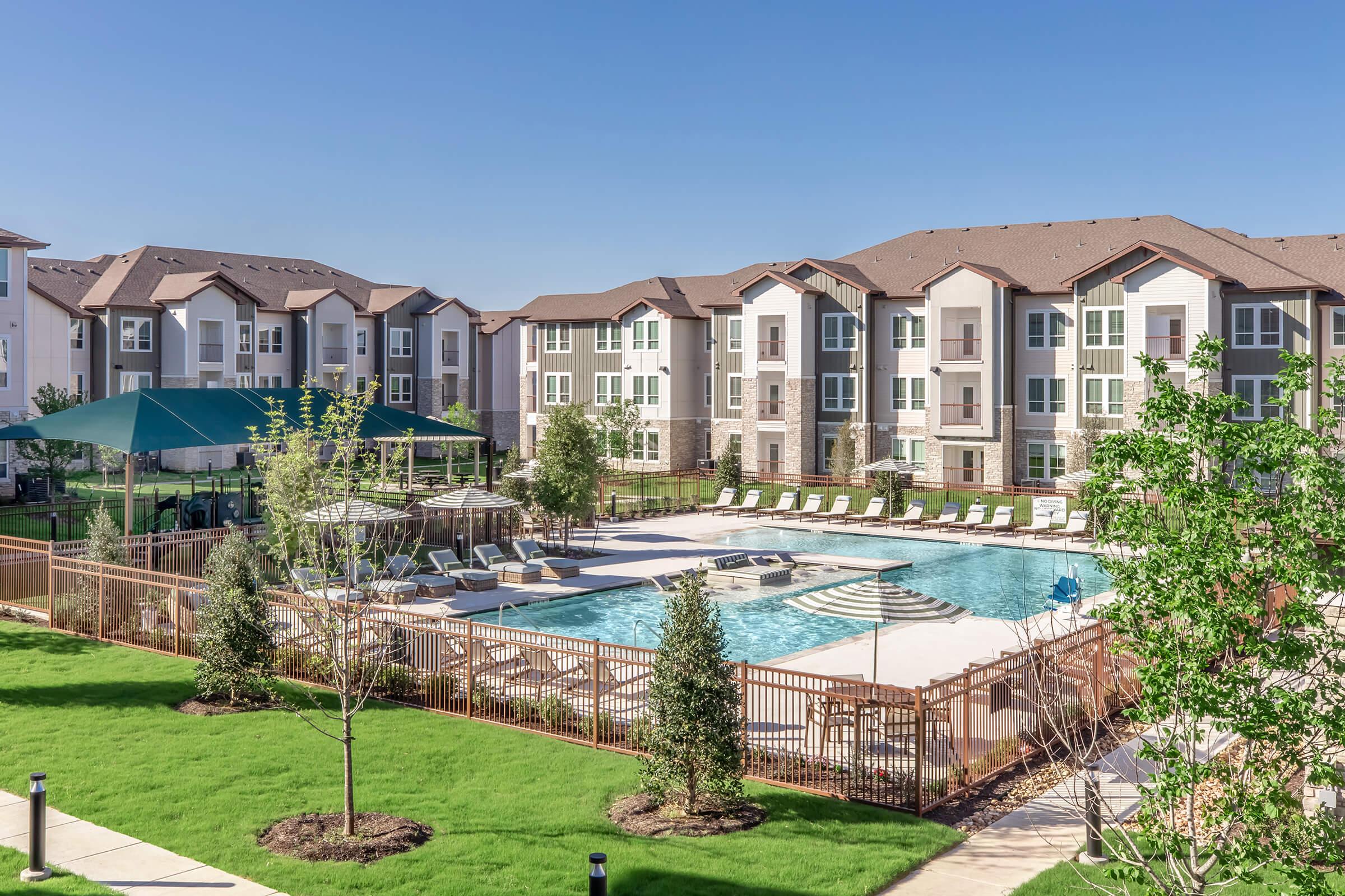 A sunny outdoor view of a modern apartment complex featuring a large swimming pool surrounded by lounge chairs. Lush green lawns and small trees are visible in the foreground. There’s a shaded seating area with a canopy near the pool. The buildings are multi-story with a contemporary design.
