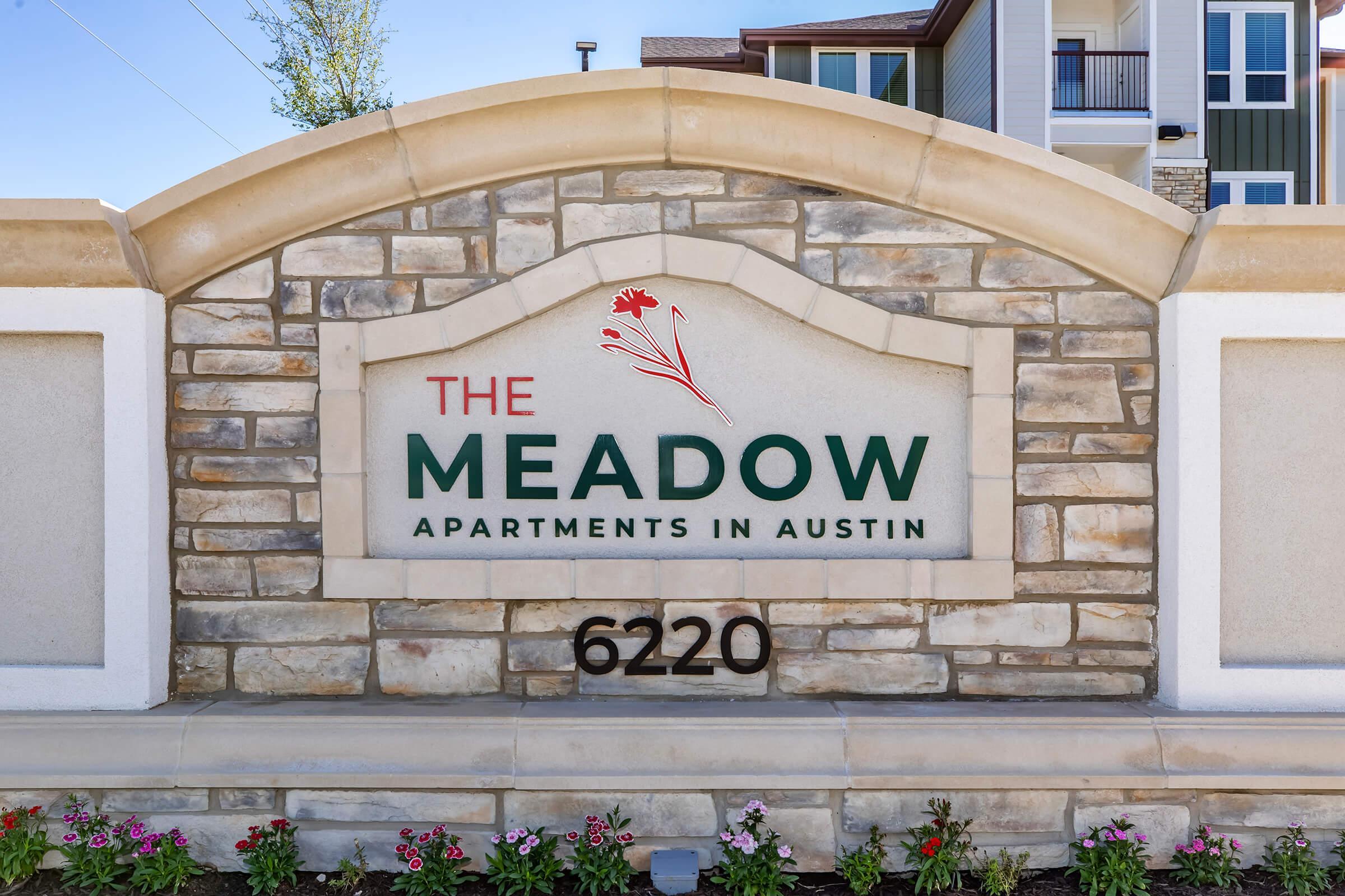 Sign for The Meadow Apartments in Austin, featuring a stone design with a floral graphic. The text displays the name "The Meadow" prominently, along with "Apartments in Austin" and the address "6220." Bright flowers are planted in front of the sign, and the background shows apartment buildings.