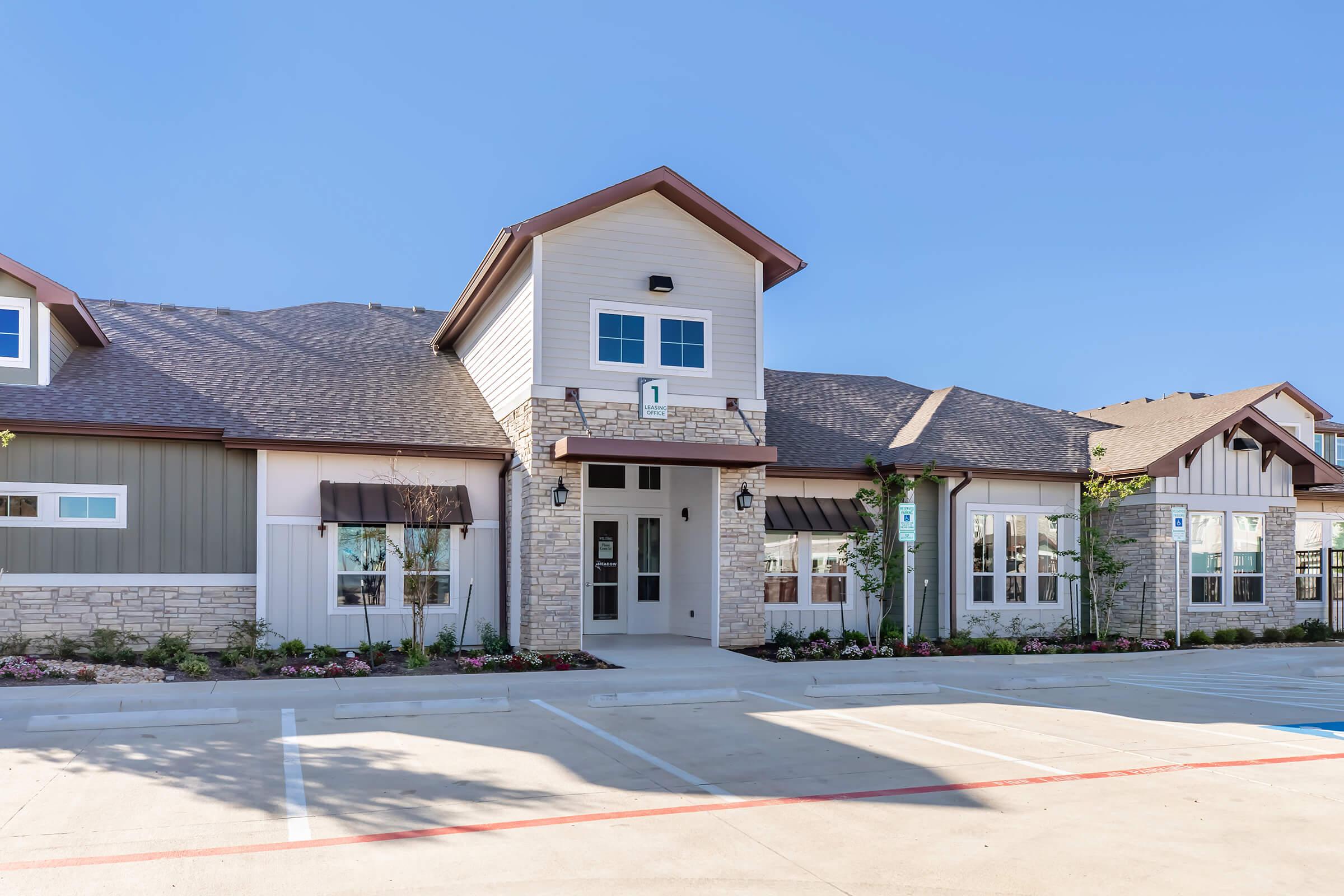 A modern one-story building with a combination of stone and siding exterior. The front features large windows and a central entrance with decorative lights. Surrounding the entrance are neatly landscaped flower beds. There are parking spaces in front of the building under a clear blue sky.
