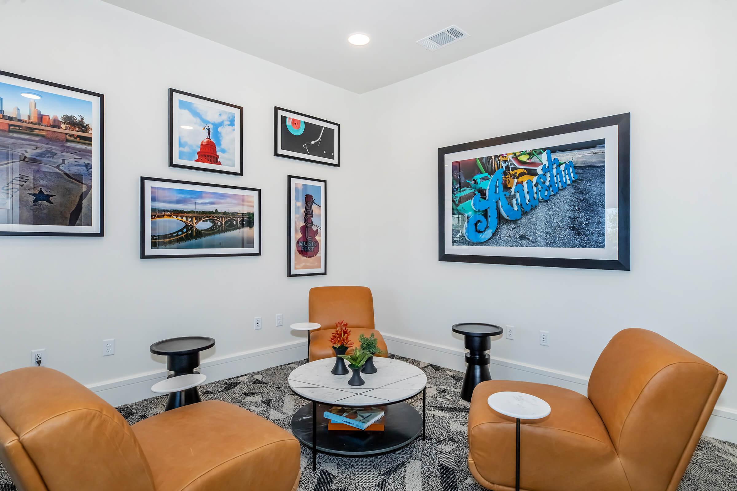 A cozy sitting area featuring two modern brown chairs and a round marble-topped table. The walls are adorned with various framed artworks, including photographs of cityscapes and colorful graffiti, enhancing the inviting atmosphere of the space.