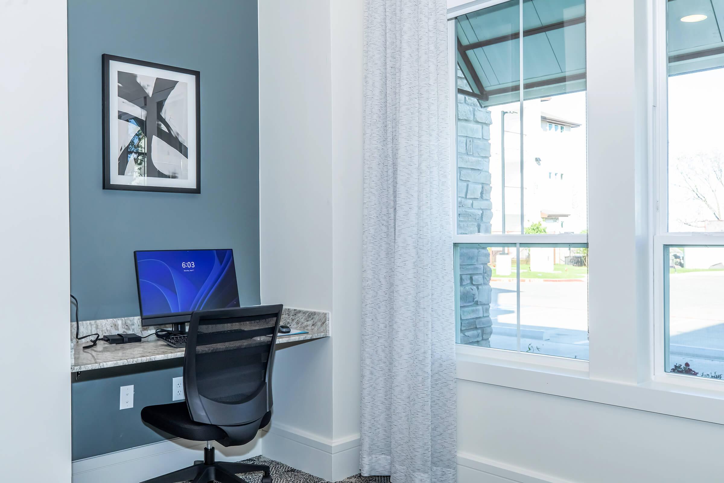 A modern home office setup featuring a computer on a grey desk, a comfortable black chair, and large windows with sheer curtains. The wall is painted in a darker shade, and a framed artwork is displayed above the desk, creating a stylish and functional workspace.