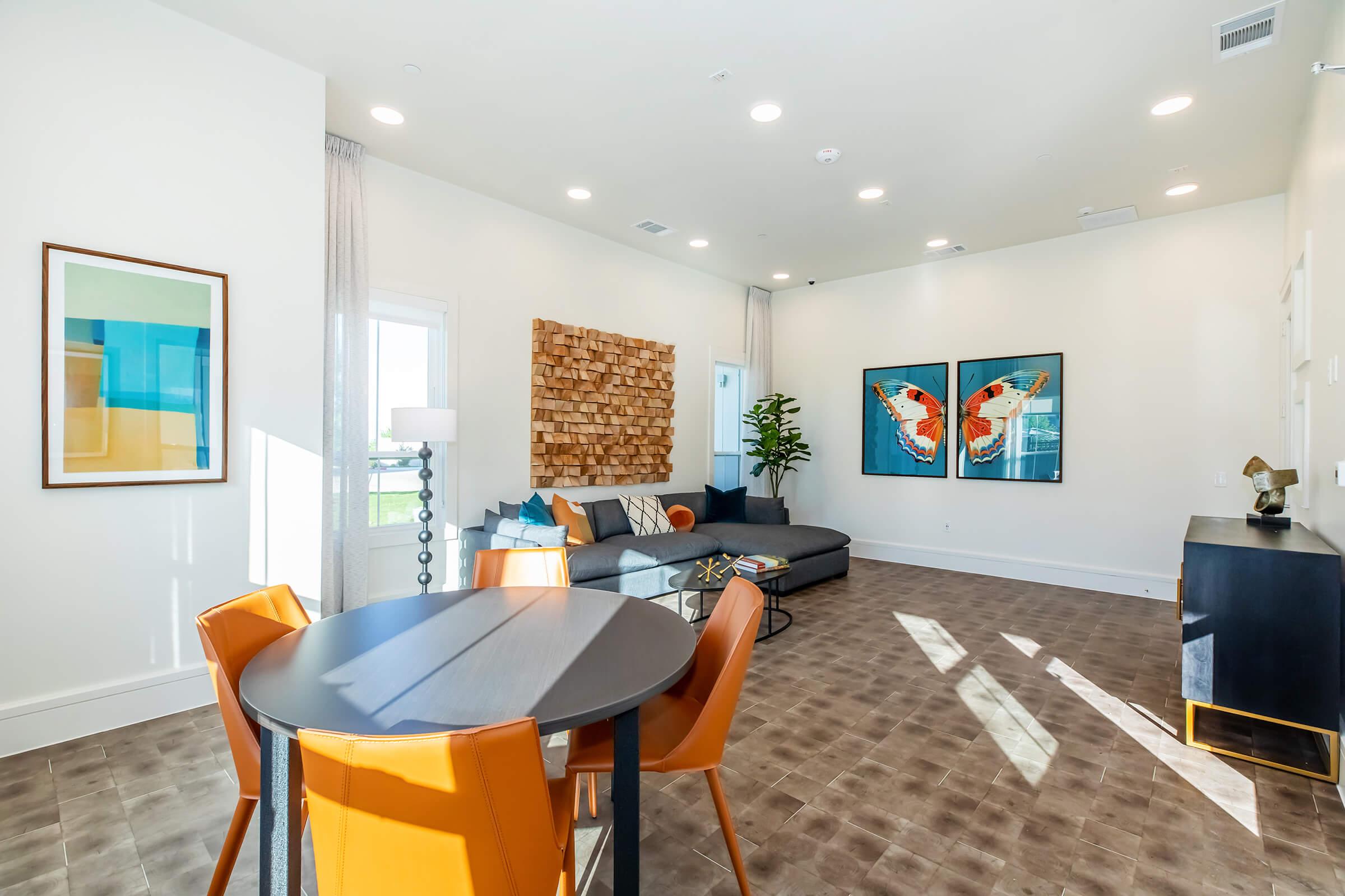 A modern living space featuring a cozy seating area with a gray couch, decorative wooden wall art, and two colorful butterfly paintings. A round dining table with orange chairs is set beside a large window, allowing natural light to fill the room. Elegant décor and a plant enhance the inviting atmosphere.
