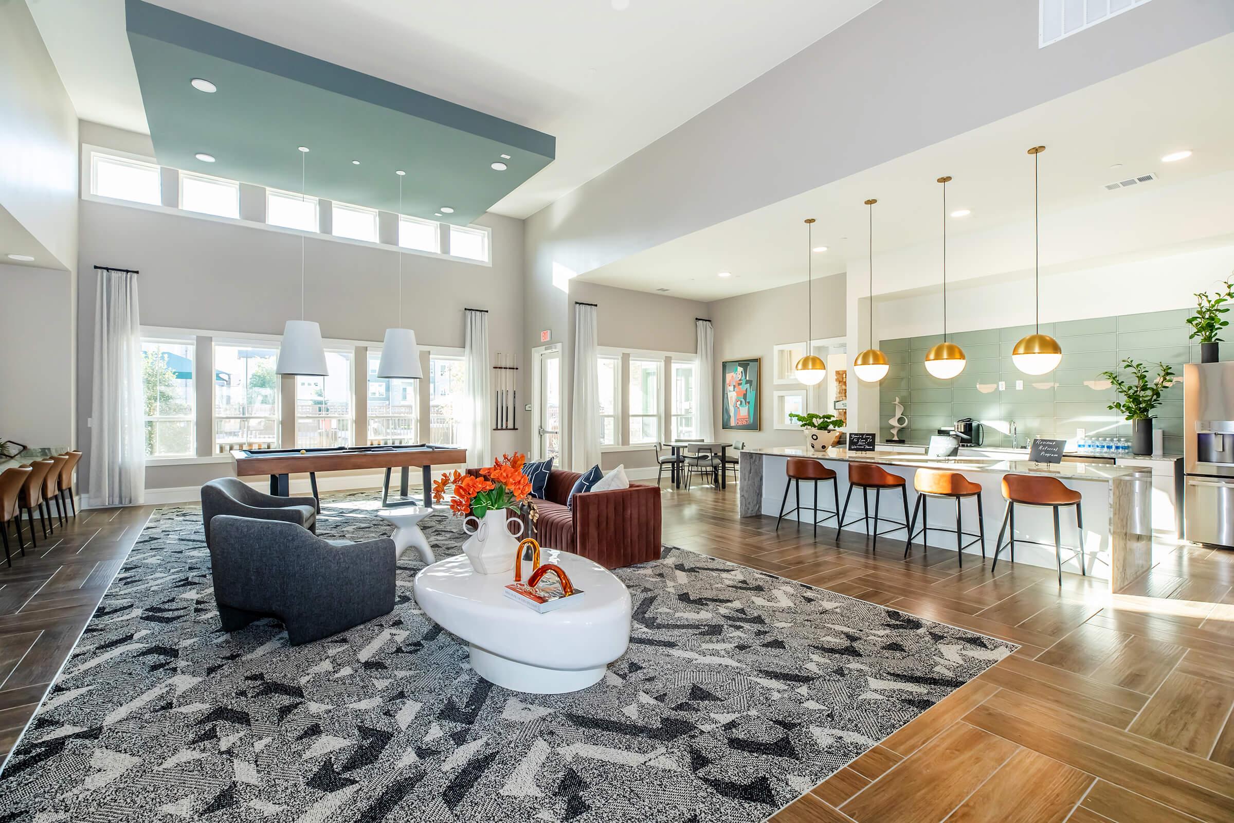 A modern community lounge featuring spacious seating areas, a large rug, and stylish finishes. It includes a dining table, comfortable chairs, pendant lighting over a kitchen island, and large windows allowing natural light to fill the space. Decorative plants and artwork enhance the contemporary atmosphere.