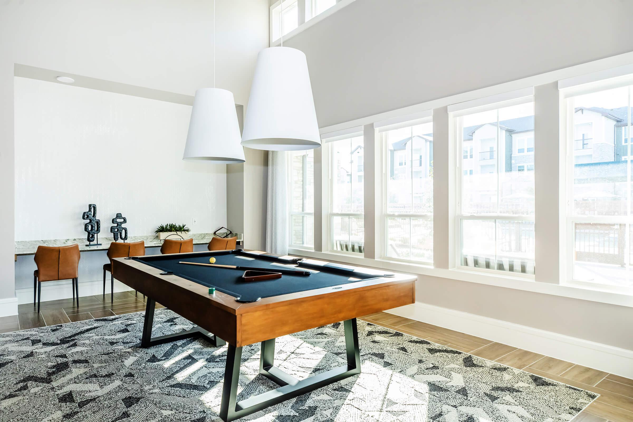 A stylish room featuring a billiard table with a dark surface, two large pendant lights overhead, and large windows allowing natural light to flow in. There is a modern dining table with two chairs and decorative items on a side table, set against a light-colored wall and patterned rug.