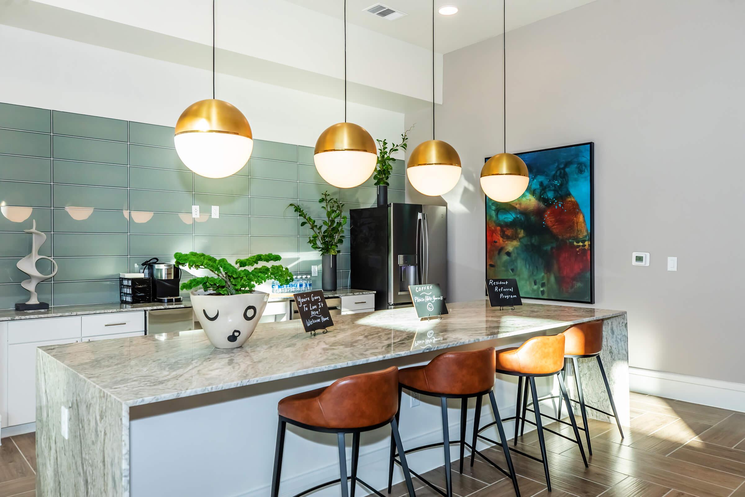 Modern kitchen featuring a spacious island with a marble countertop and four stylish brown bar stools. Above, four round pendant lights hang, illuminating the area. The kitchen includes sleek cabinetry and a black refrigerator, complemented by green plants and colorful artwork on the wall.