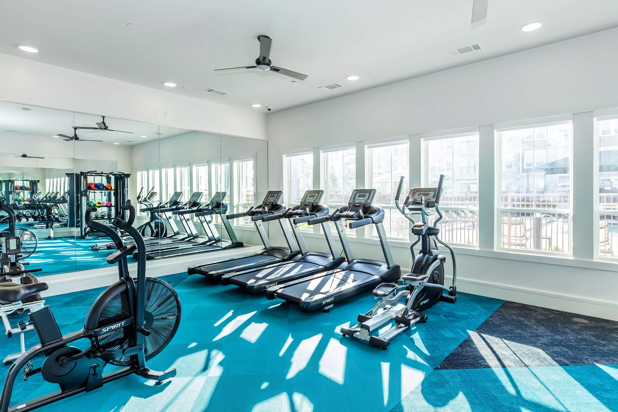 A modern gym interior featuring exercise equipment including treadmills, an elliptical machine, and a stationary bike. The room has large windows allowing natural light, a fan on the ceiling, and a blue carpet. Mirrors line one wall, creating a spacious and inviting workout environment.
