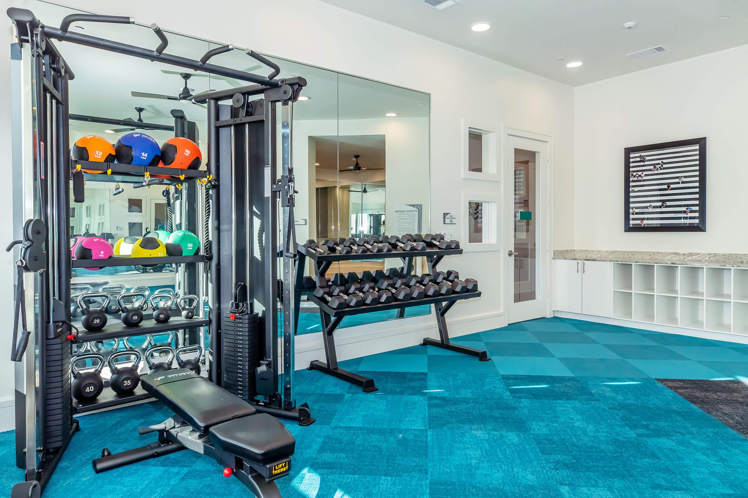 A modern gym interior featuring a weight training station with colorful dumbbells, a bench, and a rack of free weights. Mirrors line one wall, and the floor is covered in blue carpet tiles. A large whiteboard is mounted nearby, and the space is well-lit with overhead fans.