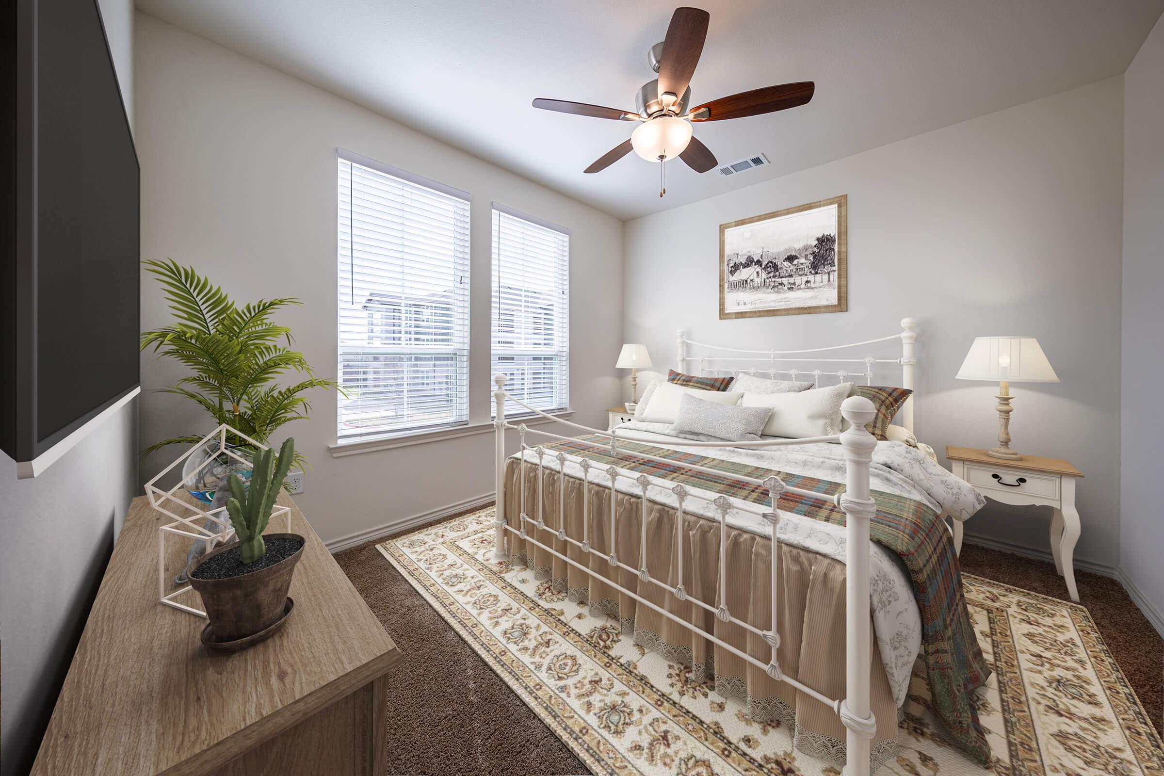 A cozy bedroom featuring a white metal bed with a patterned throw, flanked by two bedside tables with lamps. A ceiling fan is overhead, and large windows let in natural light. A potted plant and decorative items add a touch of warmth to the neutral-colored walls and carpet.
