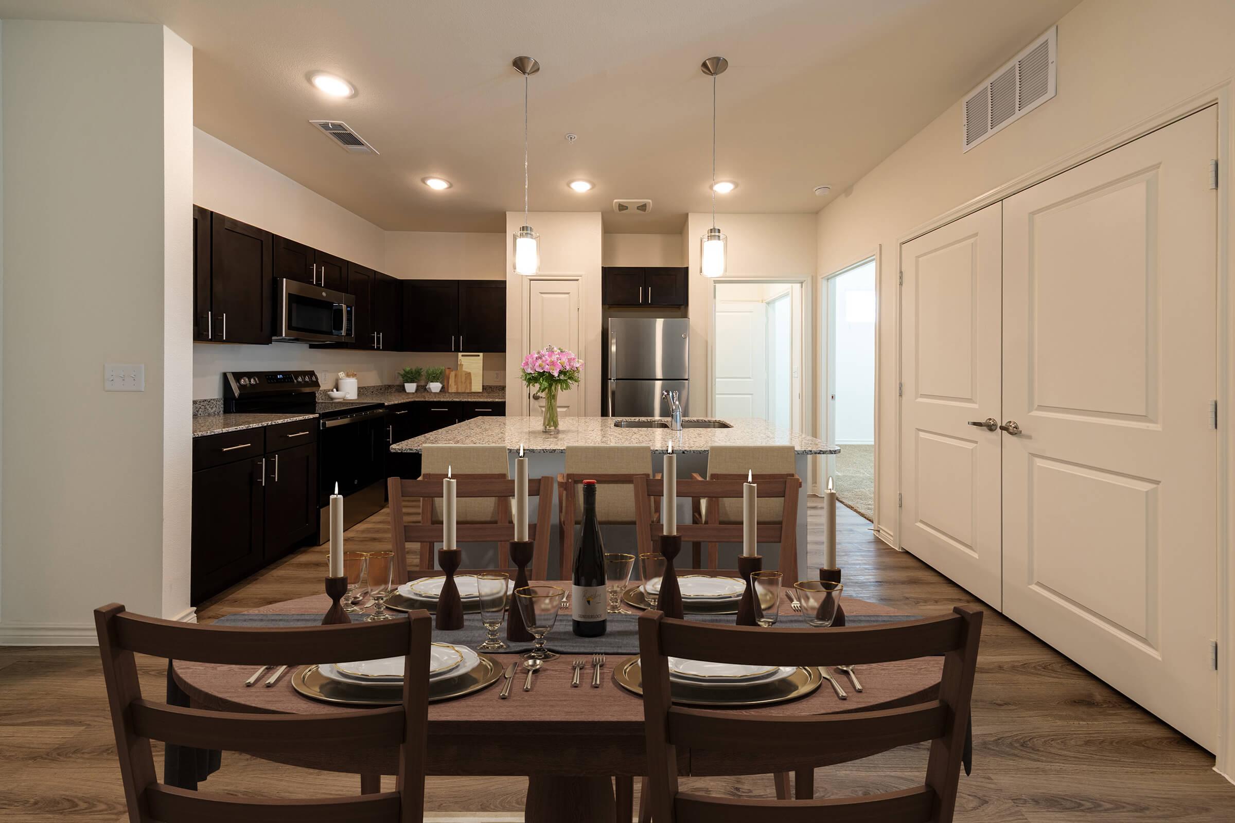 Modern kitchen and dining area featuring a dark wood dining table set for a meal with plates, glasses, and candles. The kitchen has dark cabinetry, granite countertops, stainless steel appliances, and pendant lighting. A vase with pink flowers adds color, while an open doorway leads to a well-lit room.