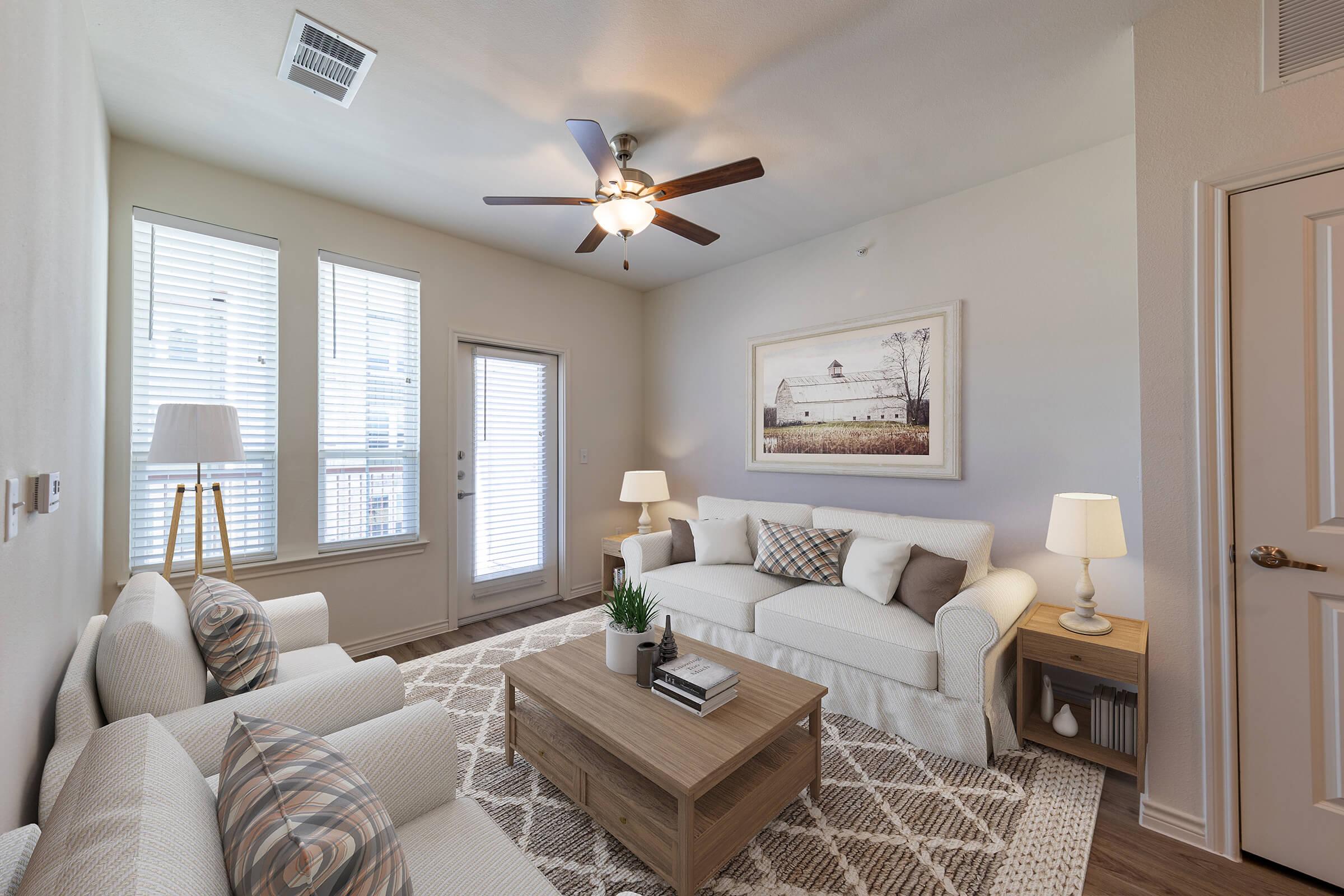 A cozy living room featuring two light-colored sofas with decorative pillows, a wooden coffee table, and a potted plant. The room has large windows with horizontal blinds, allowing natural light in. A ceiling fan and neutral-colored walls create a warm, inviting atmosphere. A framed artwork hangs on the wall.